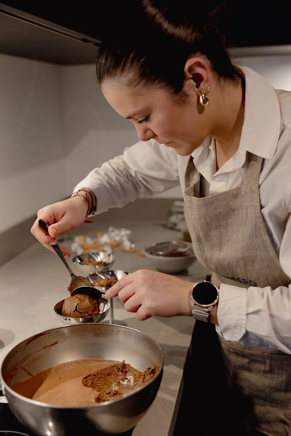 Woman wearing a beige apron and earrings, carefully serving chocolate ice cream from a scoop into small bowls in a kitchen.