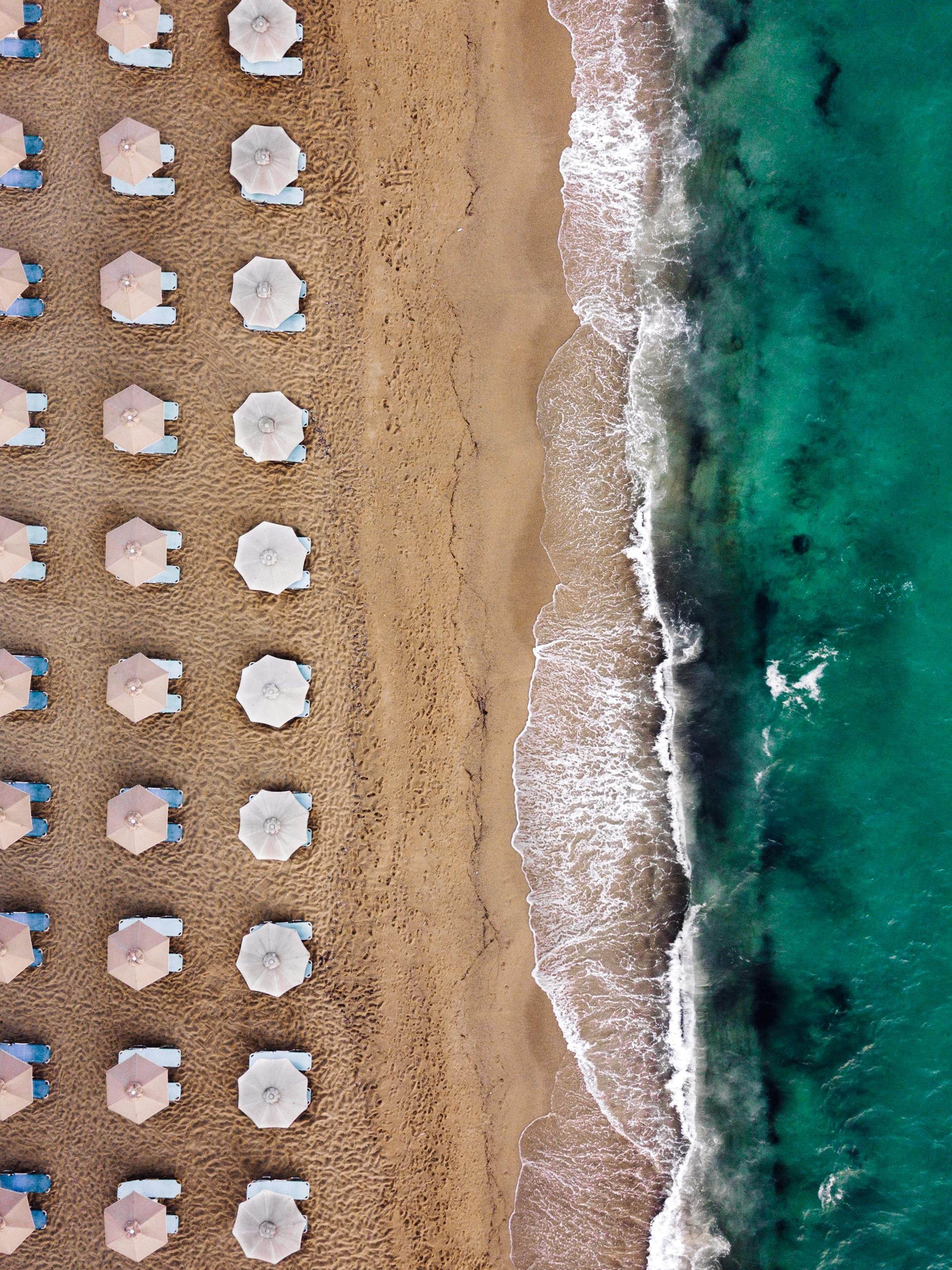 Aerial view of a beach with turquoise water, sandy shore, and a coastal town with buildings and greenery in the background.