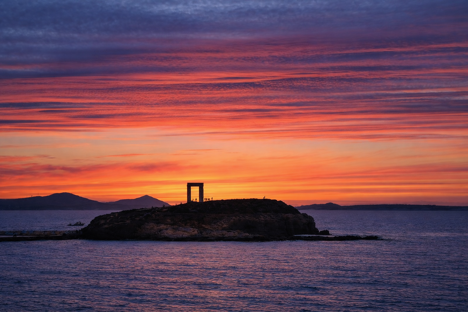 Portara of Naxos, Greece, on a hill with a clear blue sky and turquoise sea in the foreground.