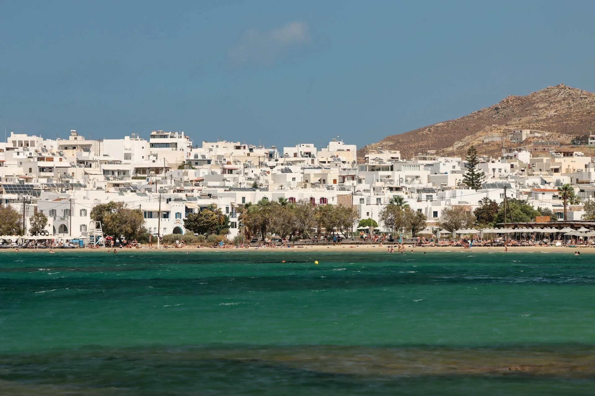 Coastal townscape with white buildings, sandy beach, turquoise sea, and hillside.