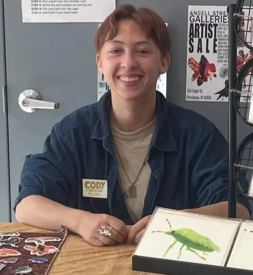 The artist, Cody Szczepanski, sits at a table. In front of him is one of his prints of a Southern Green Stink Bug.