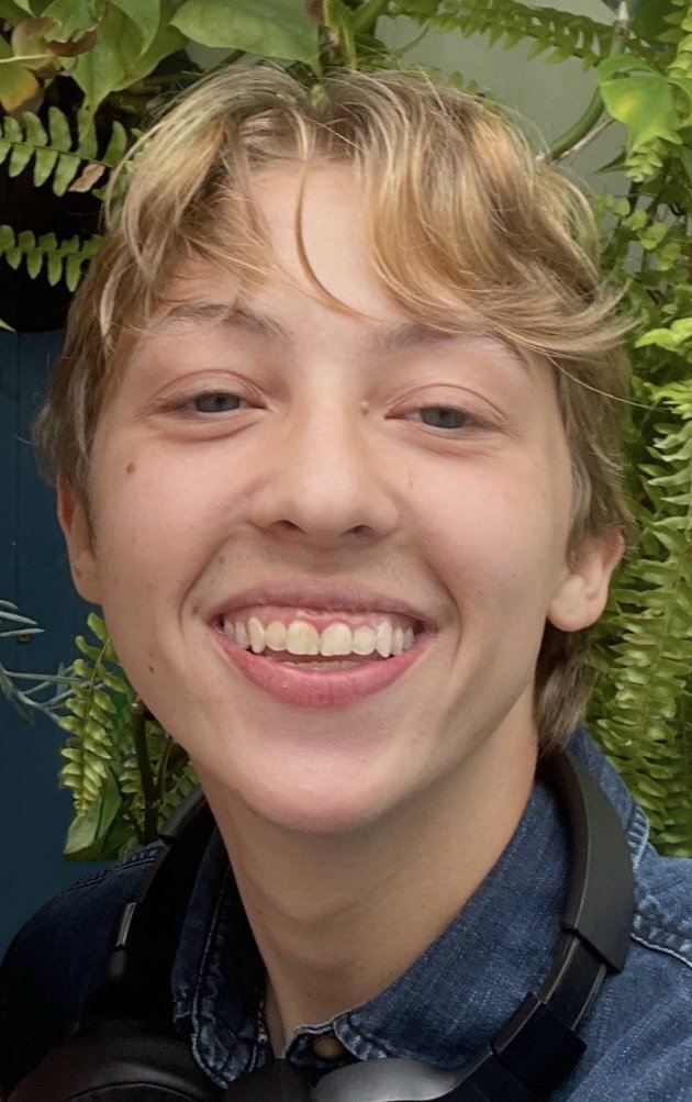 Smiling young man with light brown hair, light skin, and blue eyes, wearing a denim shirt and headphones around his neck, standing in front of green plants.