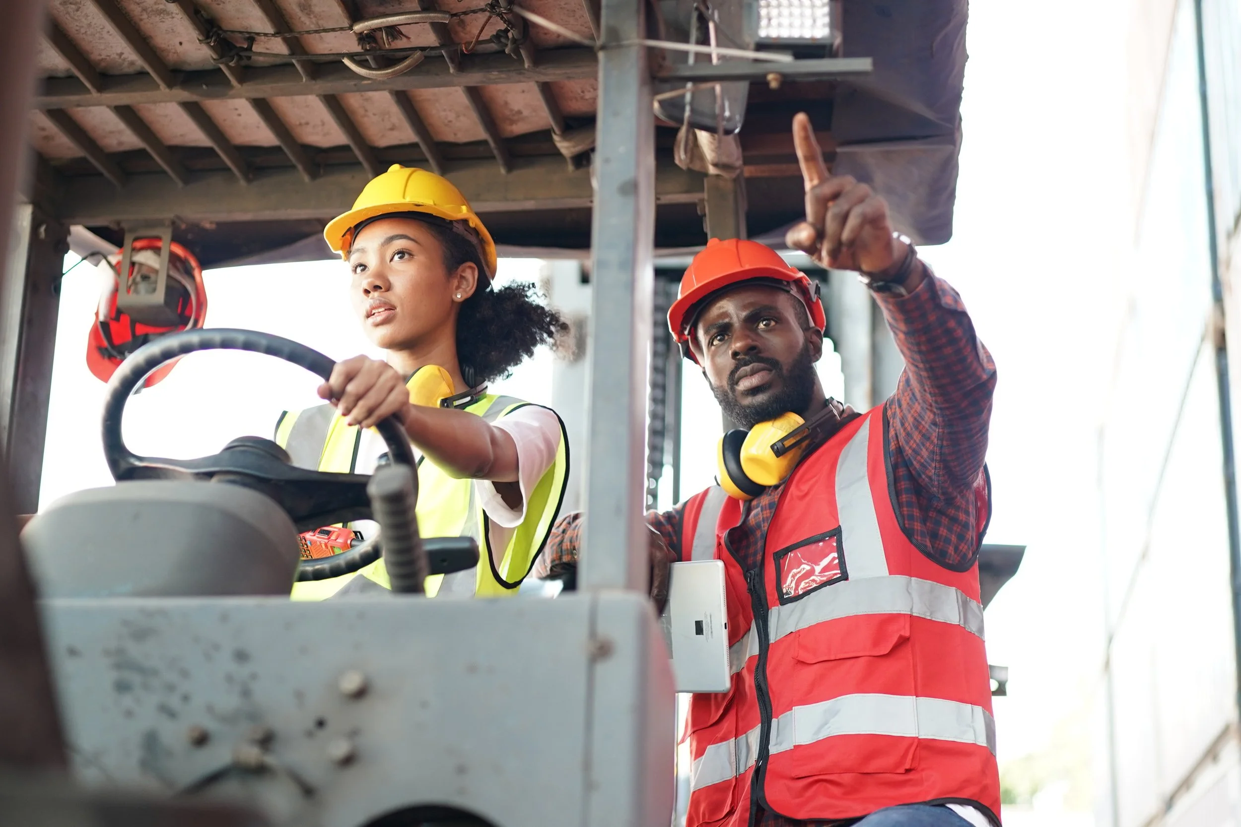 Two construction workers, a woman and a man, are on a construction site. The woman is seated in machinery, wearing a yellow hard hat and a reflective safety vest, with her hands on the controls. The man, standing next to her, is wearing an orange hard hat, a red safety vest, and has yellow ear protection around his neck. He is pointing to something in the distance, explaining or instructing.