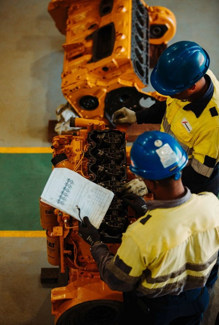 Two workers wearing safety helmets and high-visibility jackets examining and working on an orange industrial engine or machinery in a factory setting.