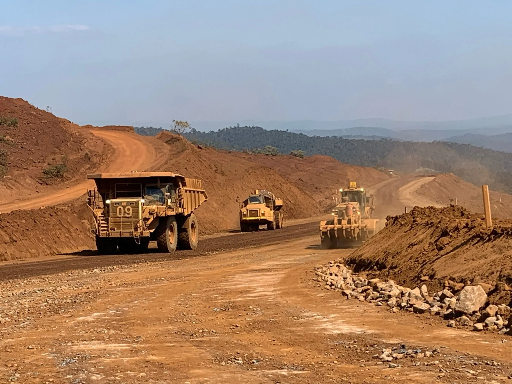 Construction site on a dirt road with heavy machinery, including a dump truck, grader, and other construction vehicles, working on leveling and paving the road in a hilly area with distant mountains.