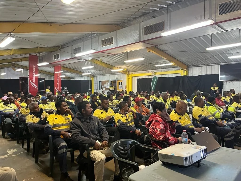 Large group of workers in yellow and black uniforms attending a presentation in a warehouse or industrial facility.