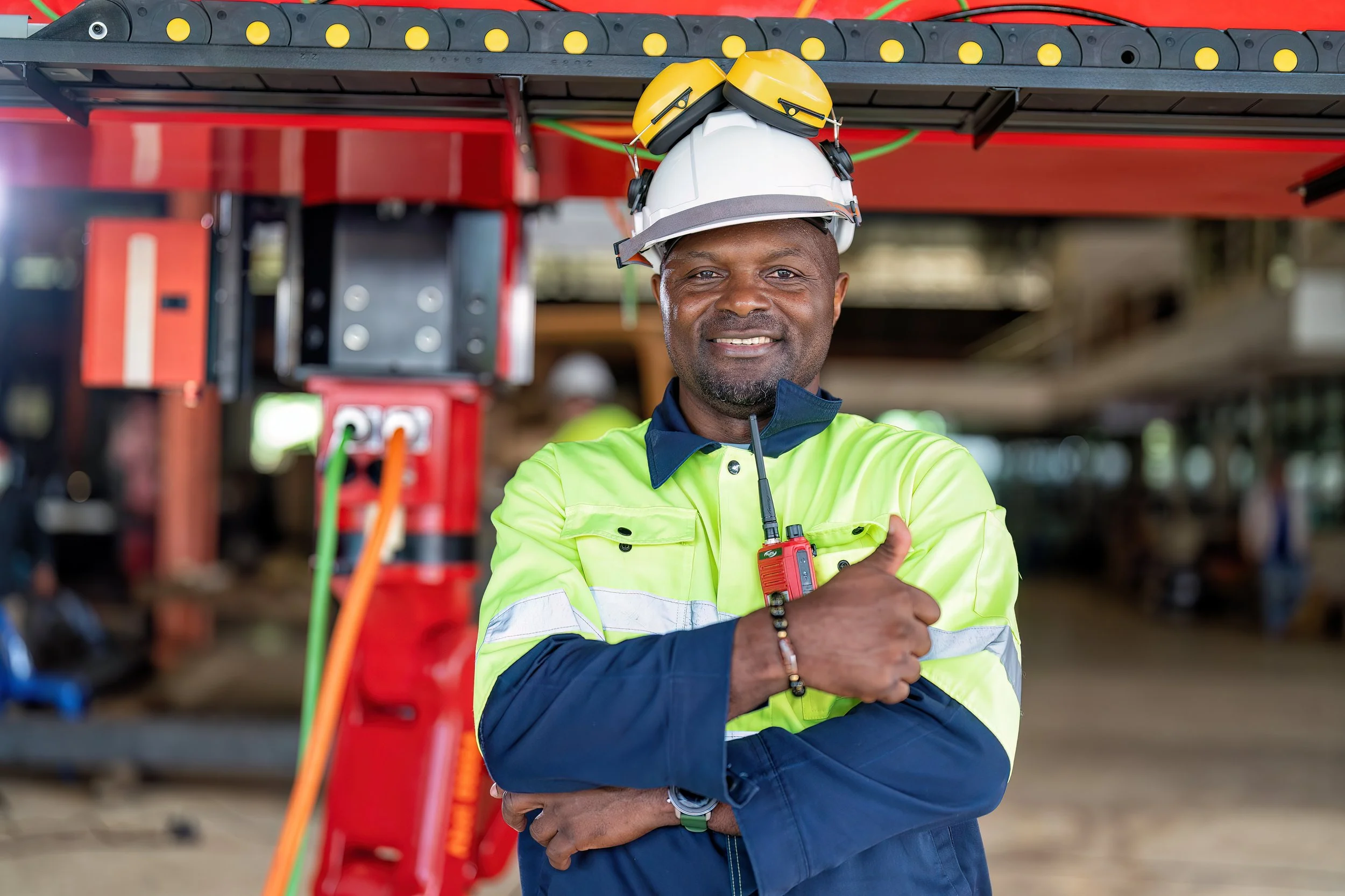 A construction worker in a yellow safety vest and a white hard hat, wearing yellow protective earmuffs, stands with crossed arms and gives a thumbs-up inside a construction site.