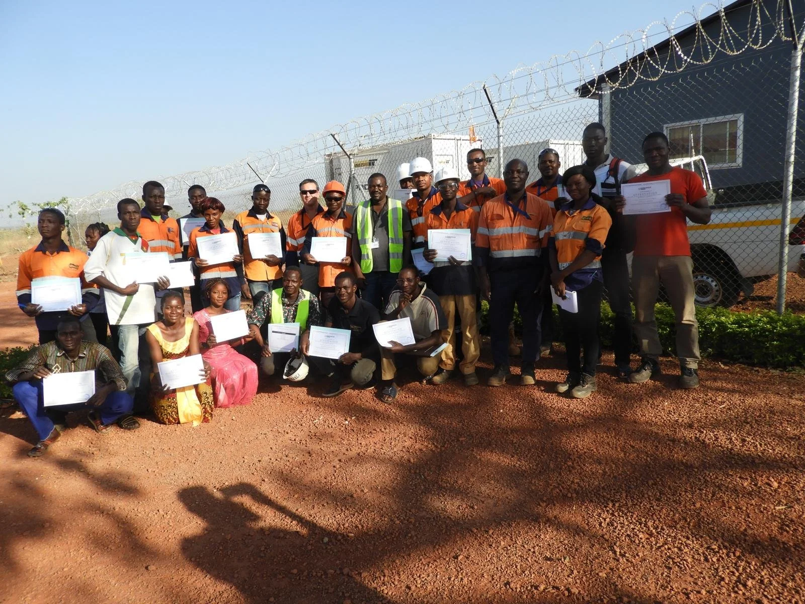 Group of people standing and kneeling outdoors near a chain-link fence, holding certificates, with some wearing safety vests and helmets, under a clear sky.