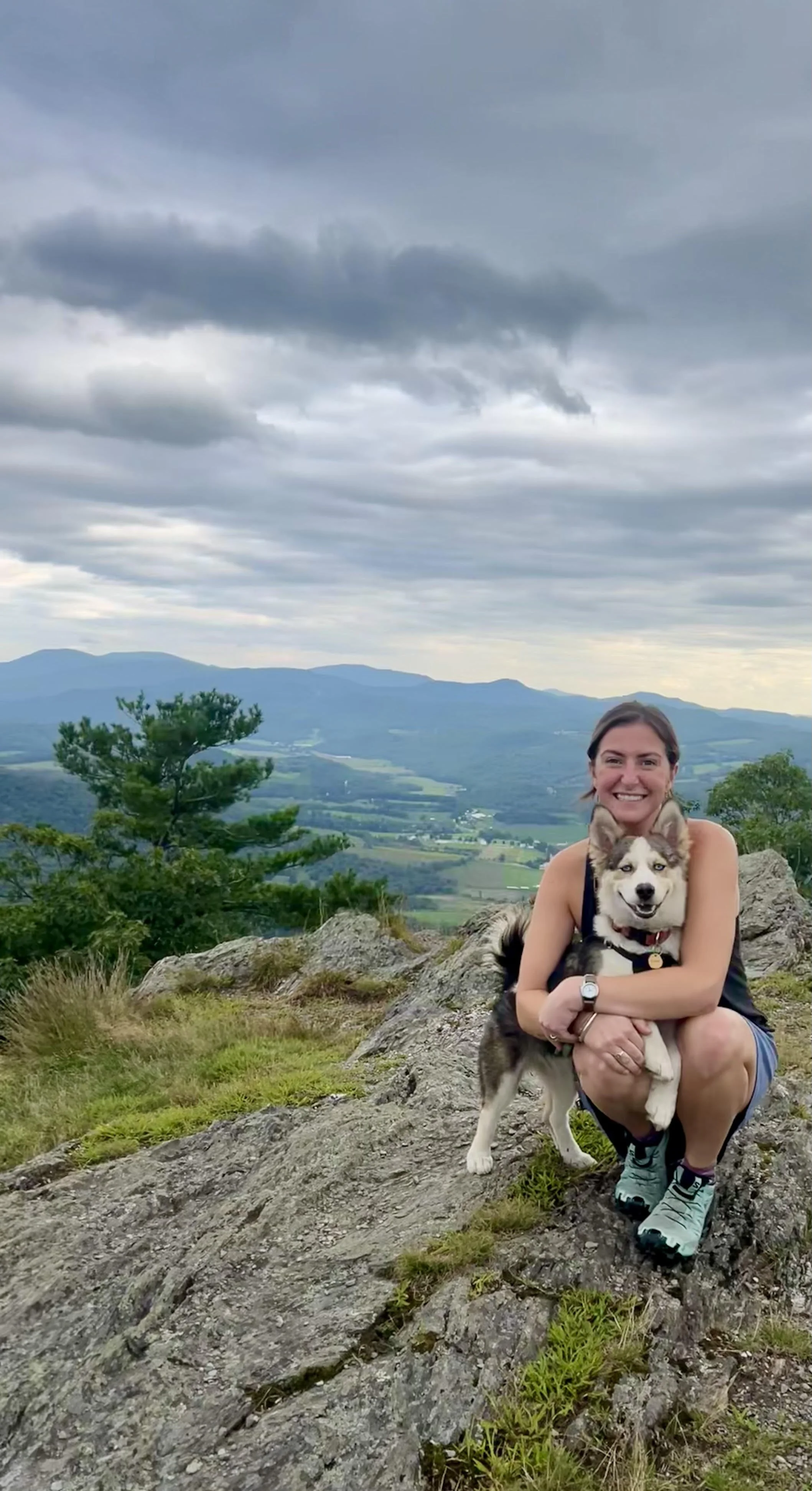 Kristin Powers sitting on a rocky hilltop with a dog, overlooking a valley with green fields and mountains under a cloudy sky.