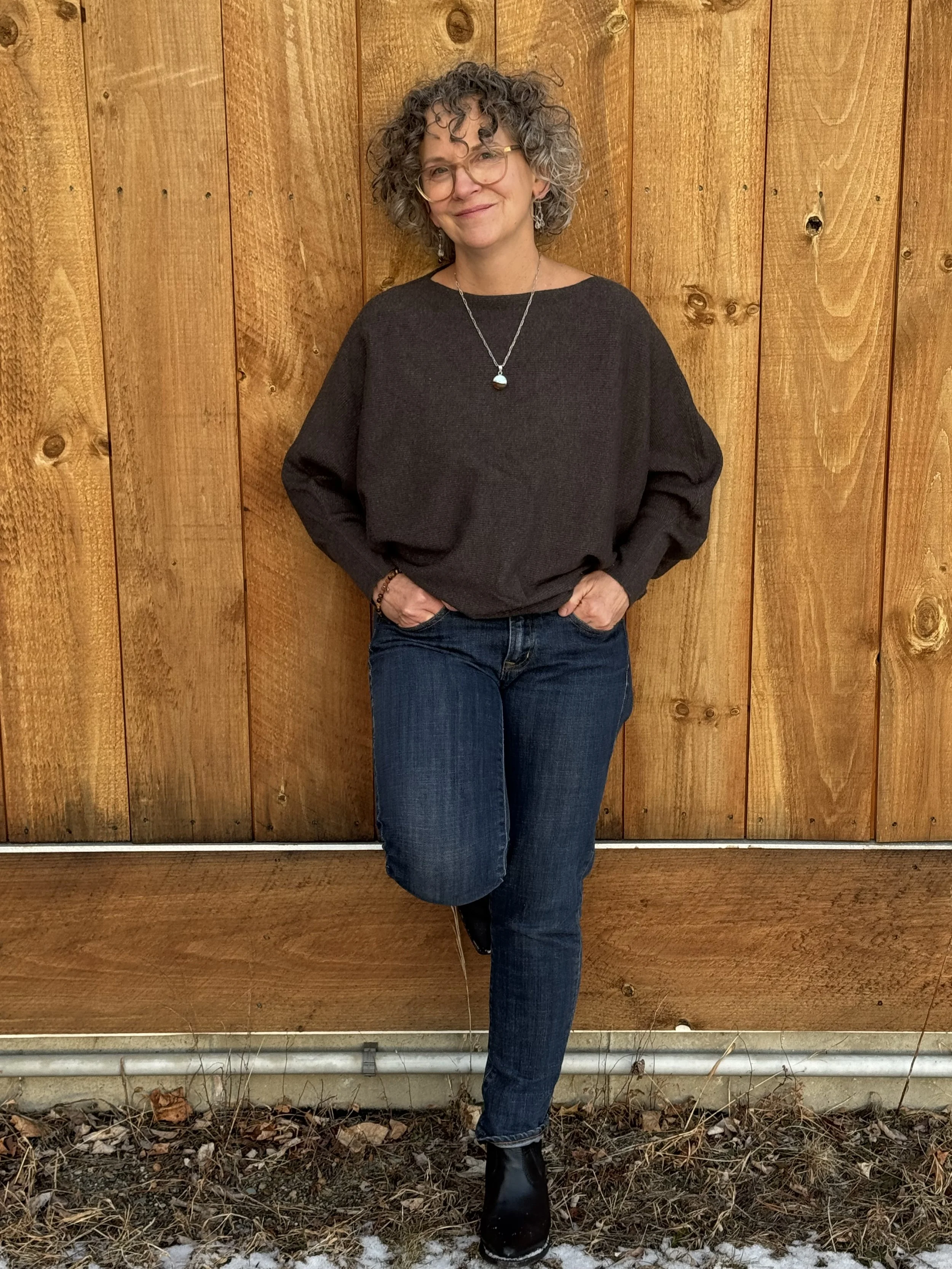 Margaret McChesney wearing glasses, a gray sweater, jeans, and black shoes, standing against a wooden wall.