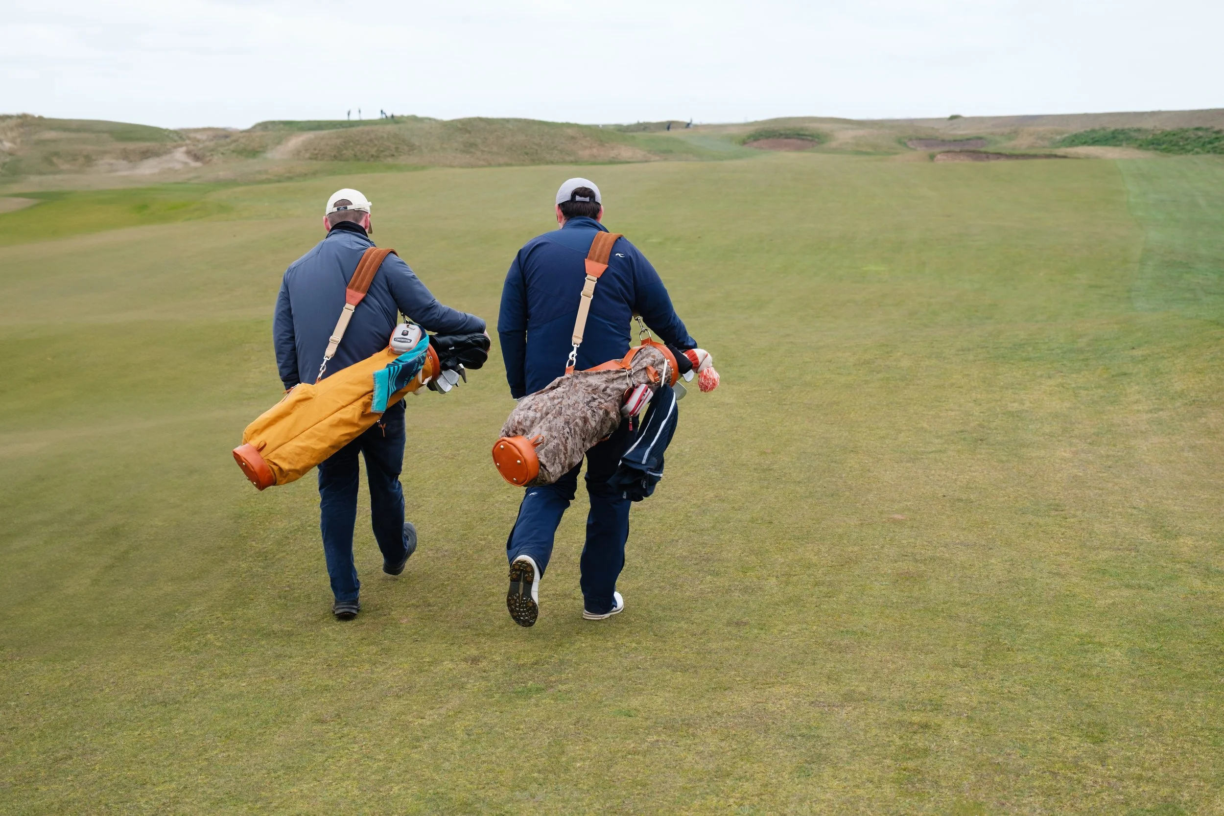 Two men carrying golf bags walk on a golf course grassy fairway with a hilly landscape in the background.