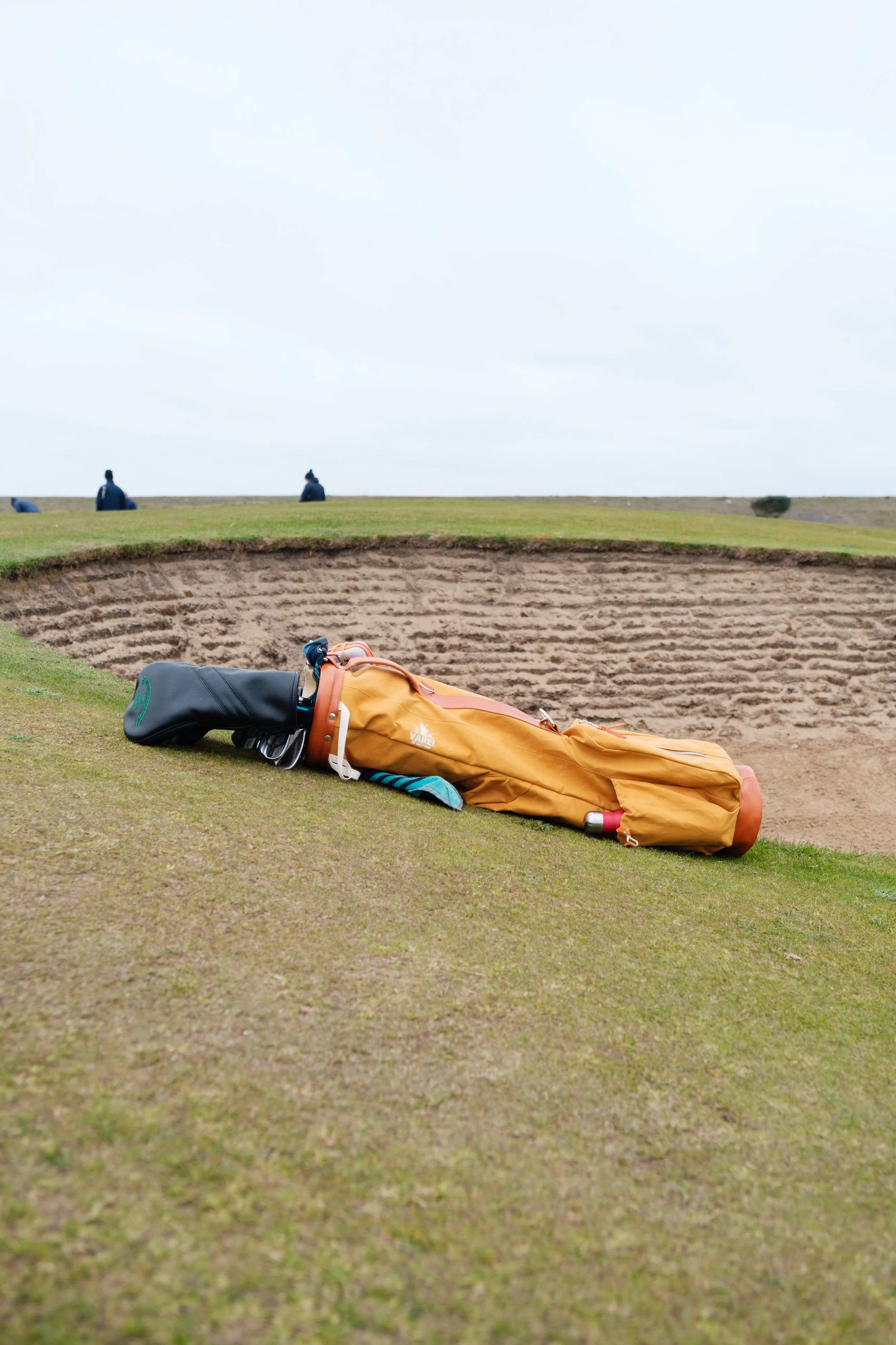 A golf bag lying on the edge of a golf green near a sand trap, with a sand wedge club visible, and people sitting on a hill in the background under an overcast sky.