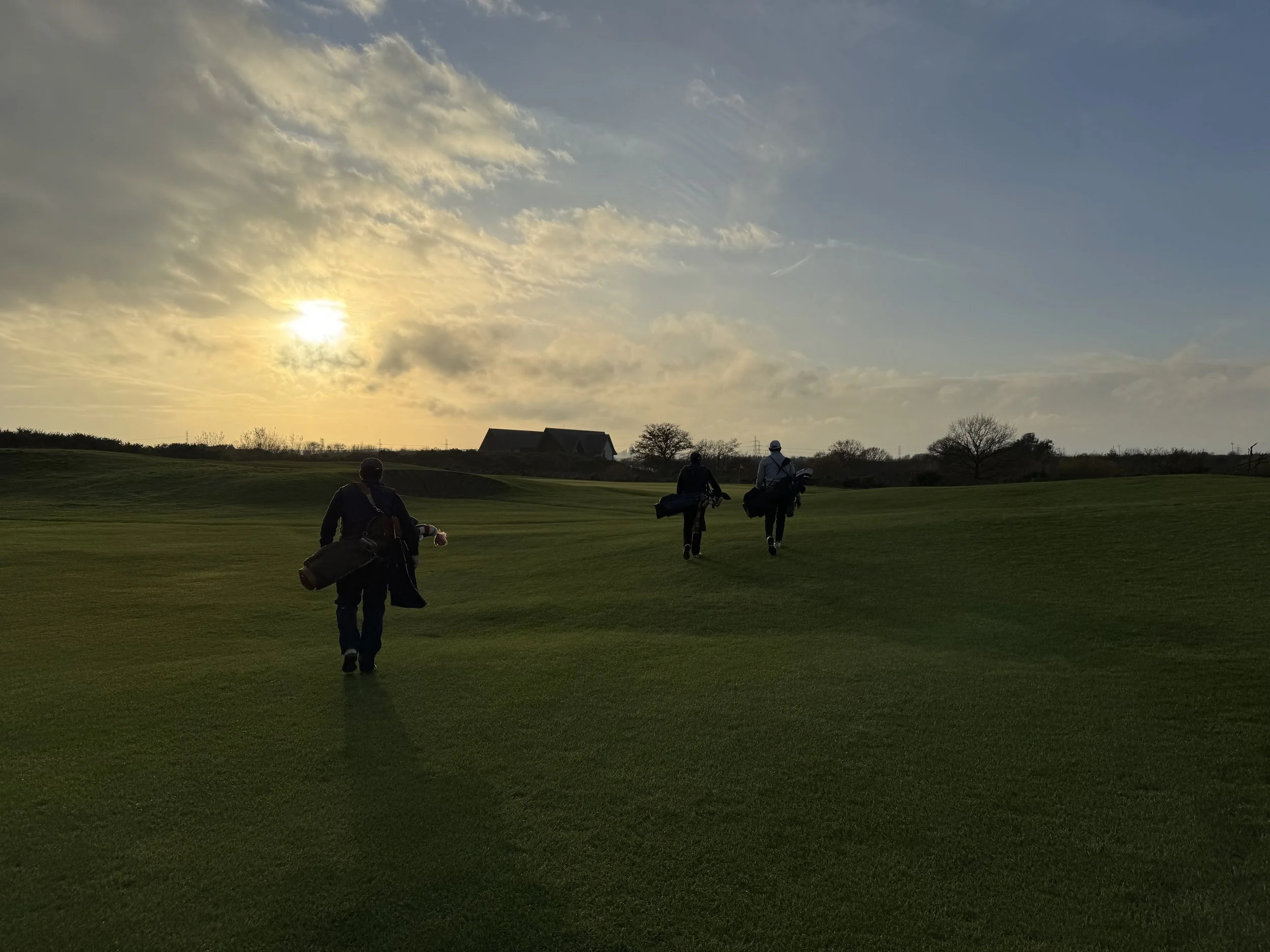 Three golfers walking across a golf course during sunset, each carrying golf bags.