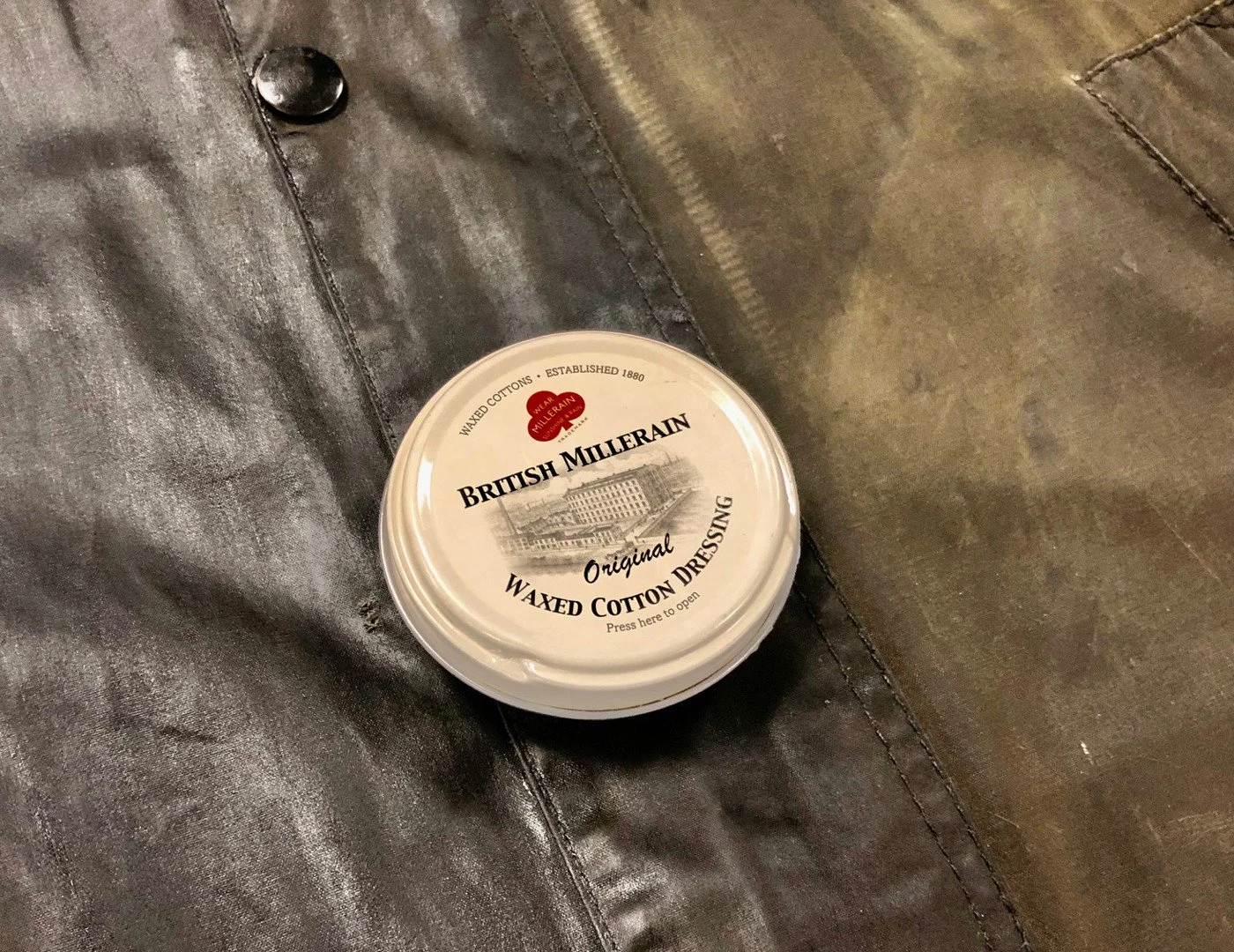 A round container of British Millerain waxed cotton dressing resting on a leather surface.