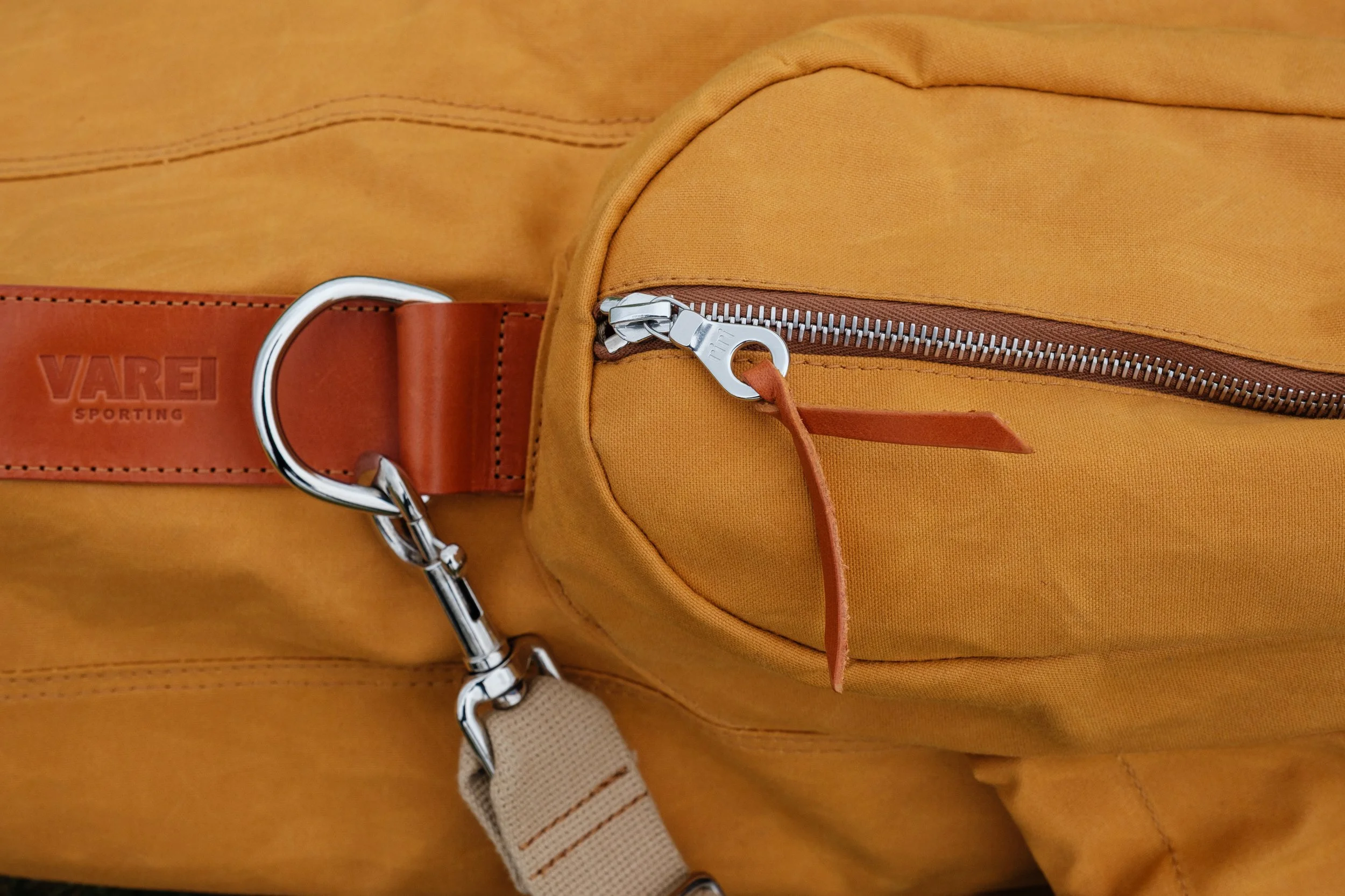 Close-up of an orange bag with a zipped pocket, a brown leather strap. The strap has the word 'VAREI' and 'SPORTING' embossed on it, and a metal clasp clip.