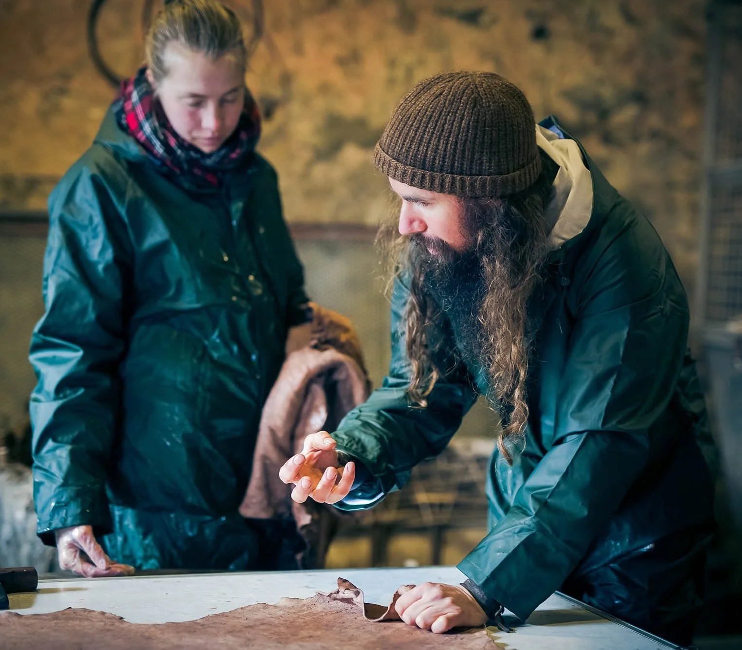 A man with long hair and beard in a winter jacket and beanie explaining or examining an object to a woman in a winter coat and scarf during daytime indoors.