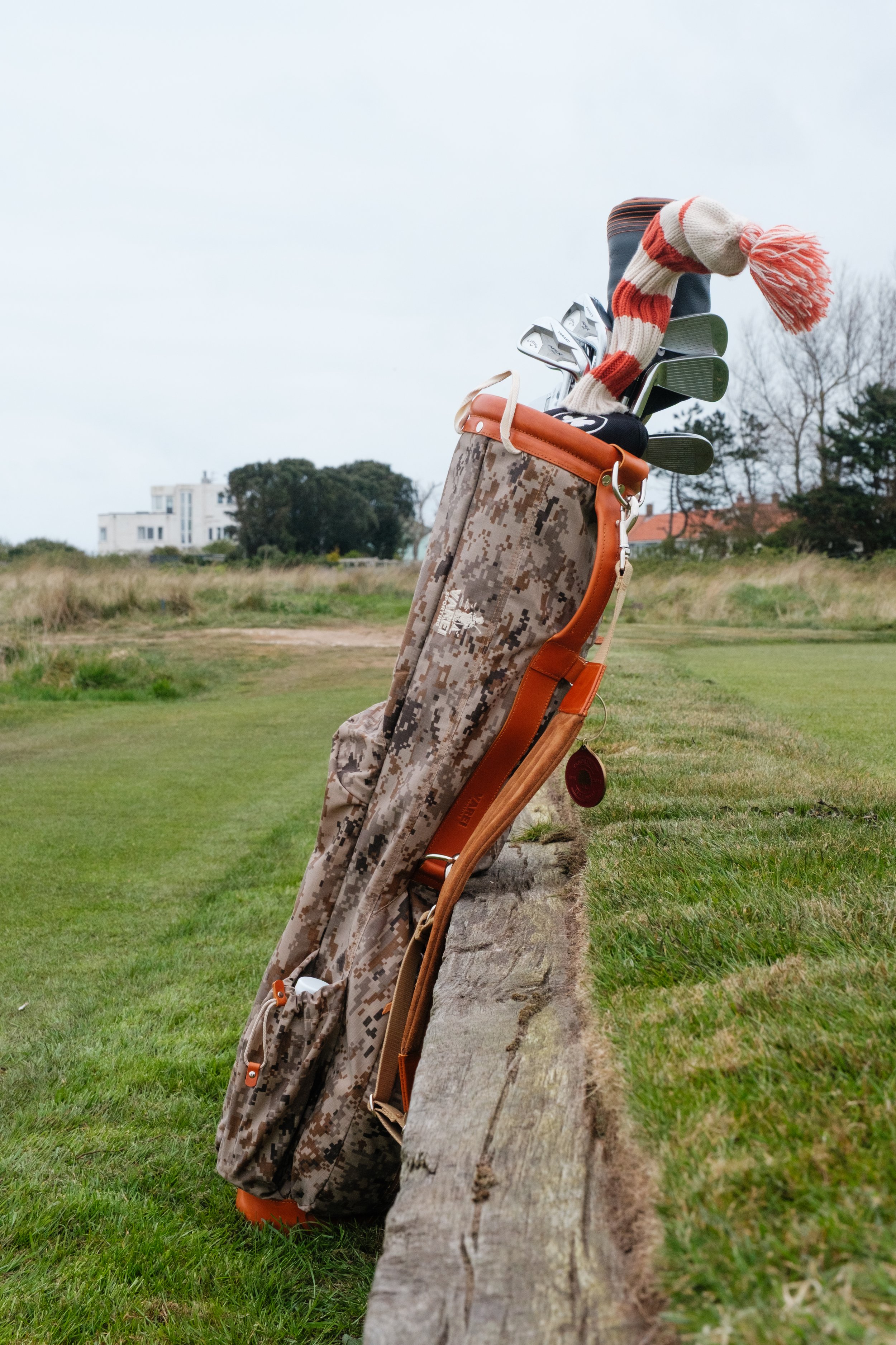 Golf bag with clubs and a red and white striped knit cap leaning against a wooden barrier on a golf course.