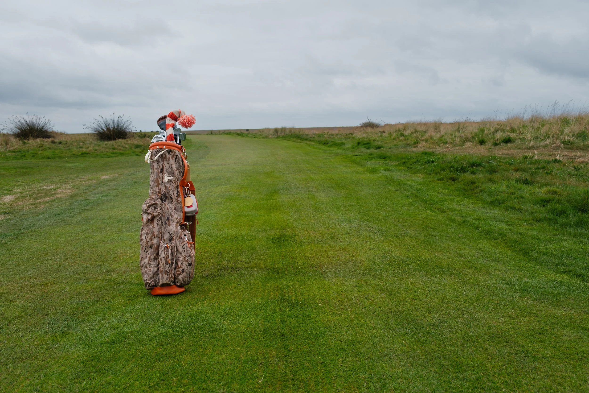 A golf bag with clubs and a red and white striped cap on a golf course with green grass and a cloudy sky.