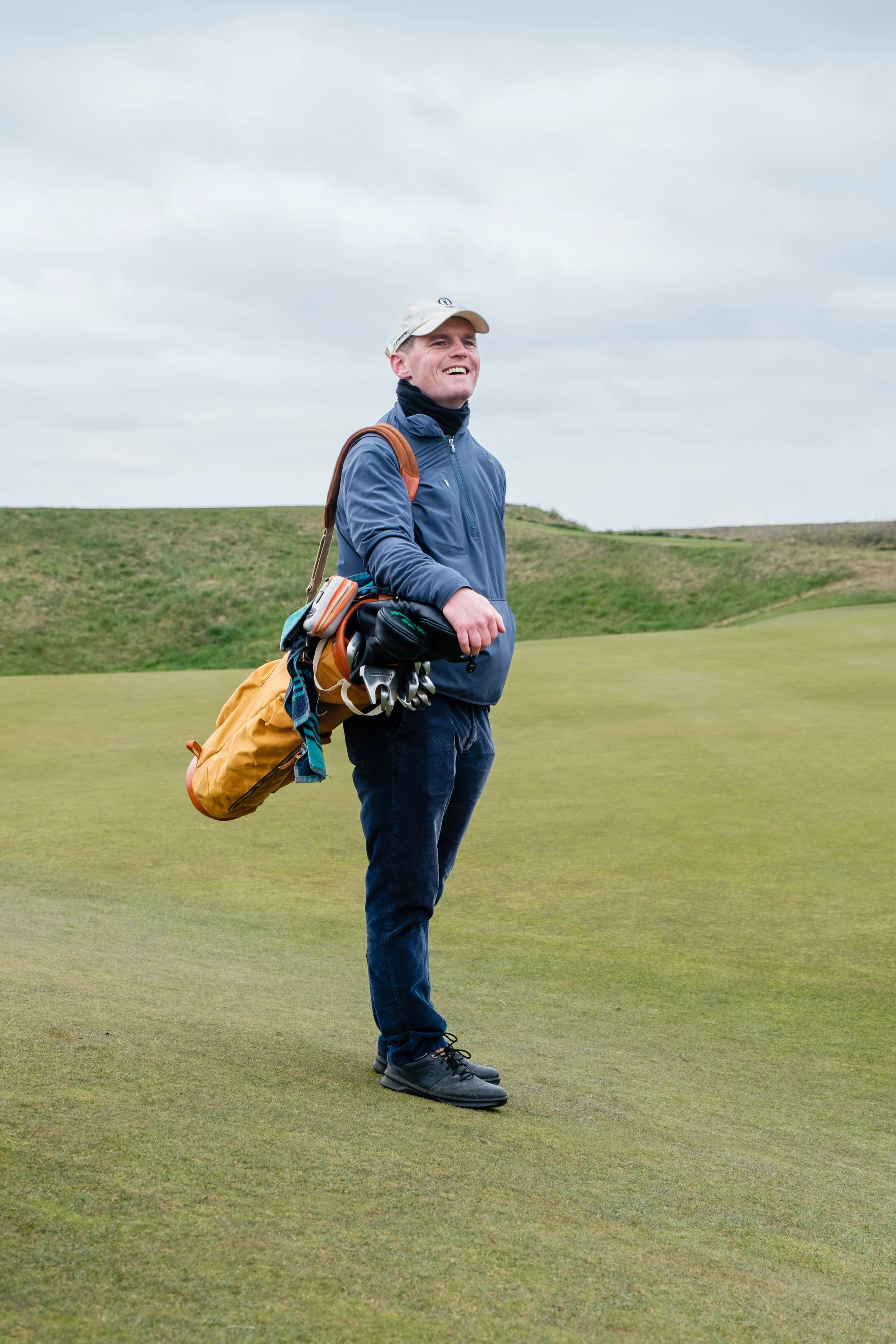 A man standing on a golf course, smiling and looking into the distance, dressed in a blue jacket, dark pants, and a baseball cap, with a golf bag filled with clubs slung over his shoulder.