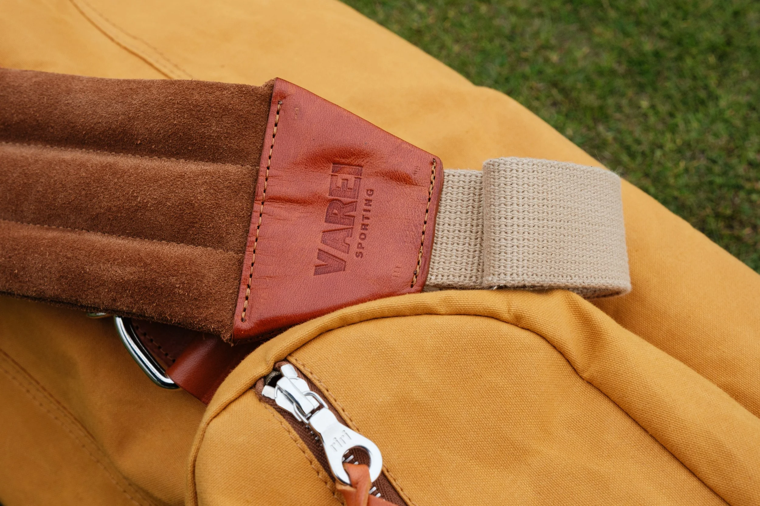 Close-up of a Varde Sporting belt with a brown suede strap, leather accents, and a beige elastic strap, placed on a yellow bag with a zipper, outdoors on grass.