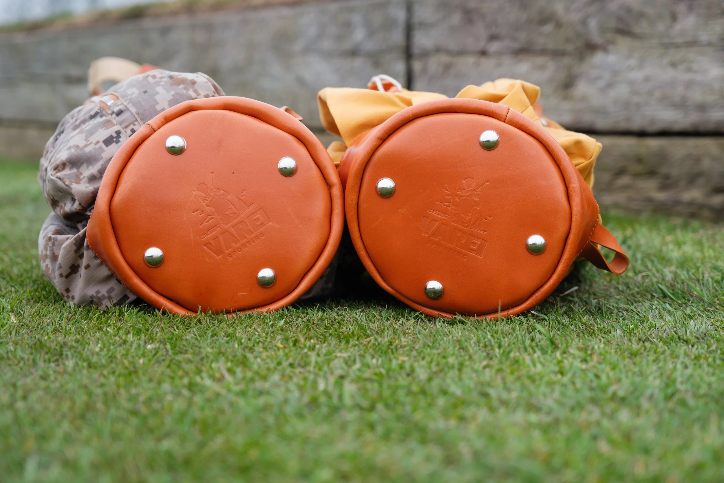 Two orange sports bags with silver studs on the bottom, placed on grass, with a wooden background.