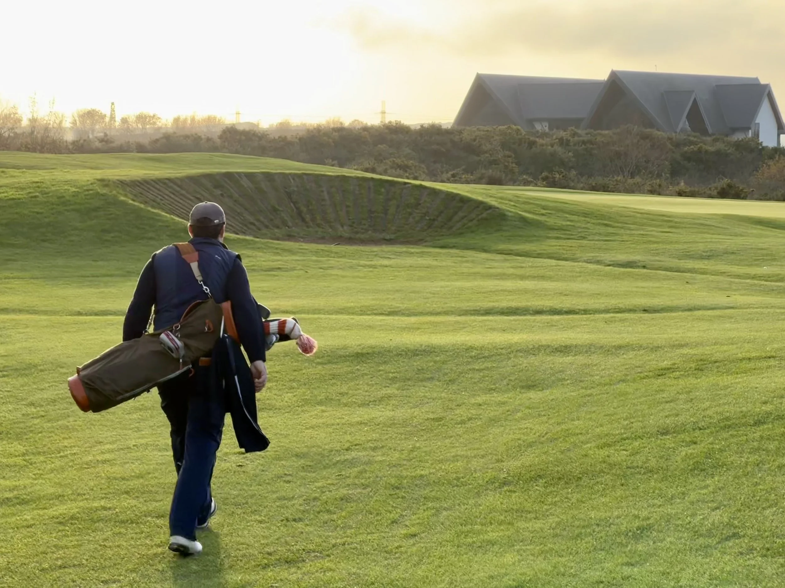 A man walking on a golf course with a golf bag over his shoulder, heading toward a sand trap, with houses in the background during sunset.