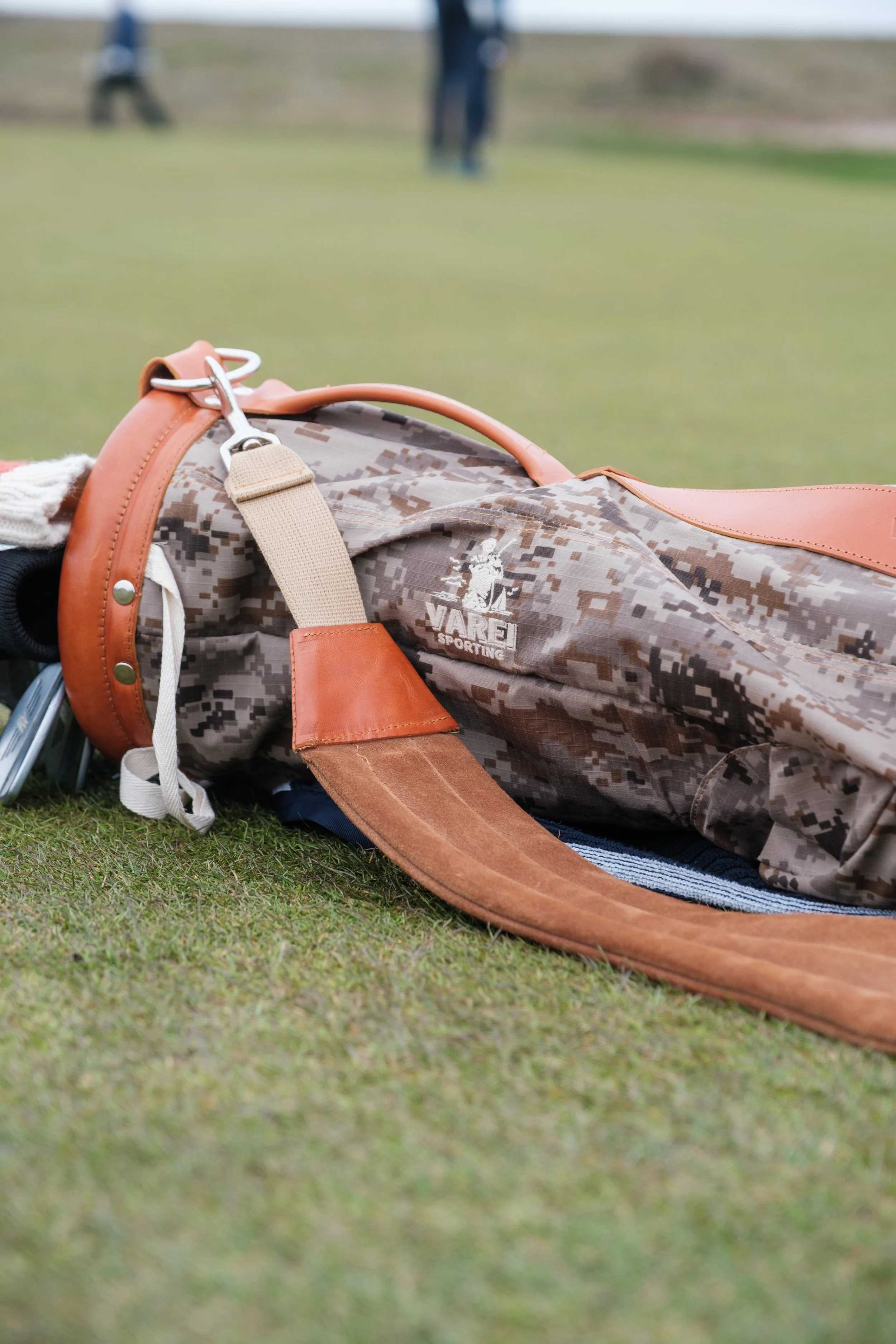A camouflage-patterned golf bag lying on the grass at a golf course with a blurred background of people and trees.