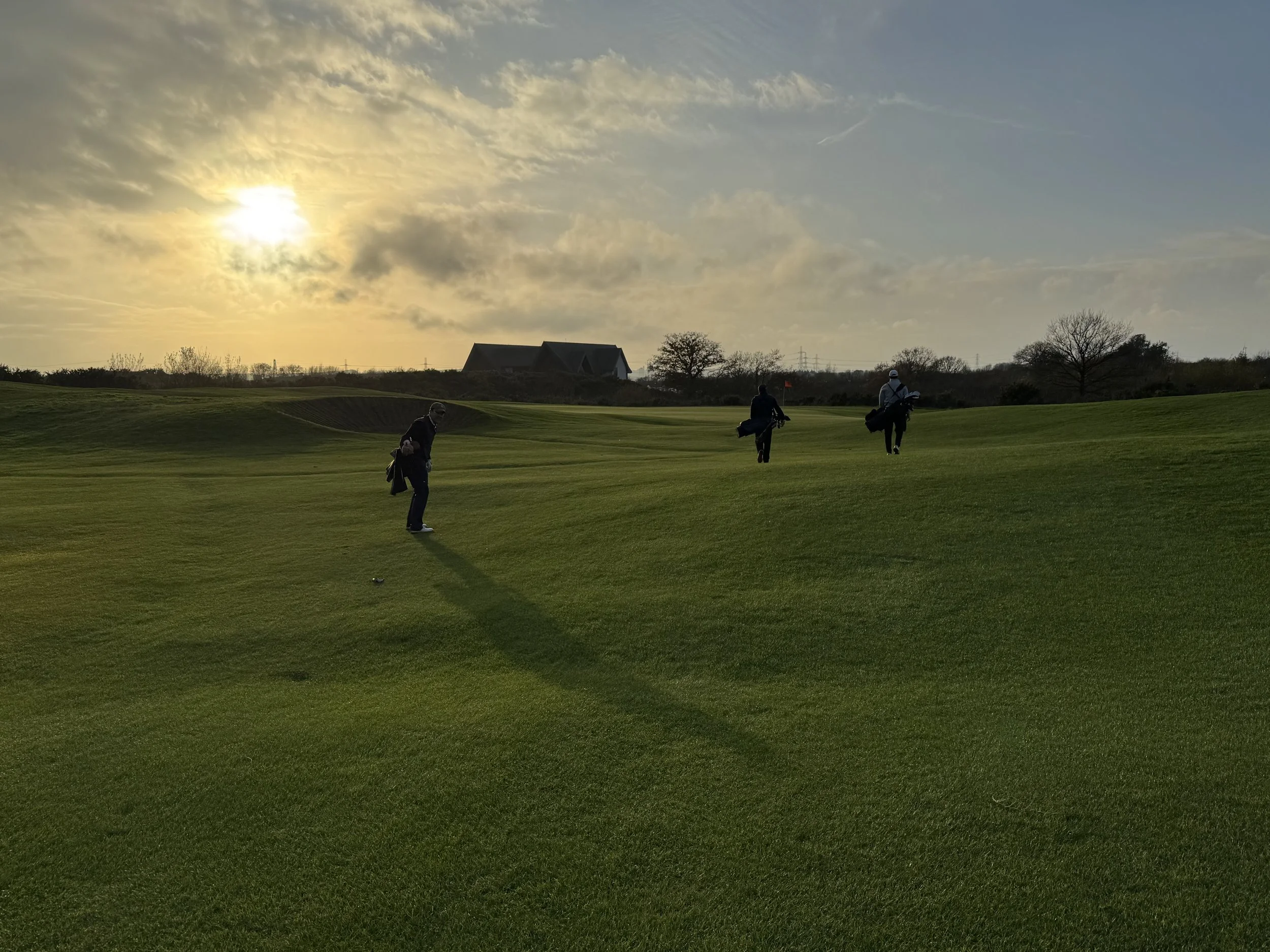 Three golfers walking on a golf course during sunset, with a house and trees in the background.