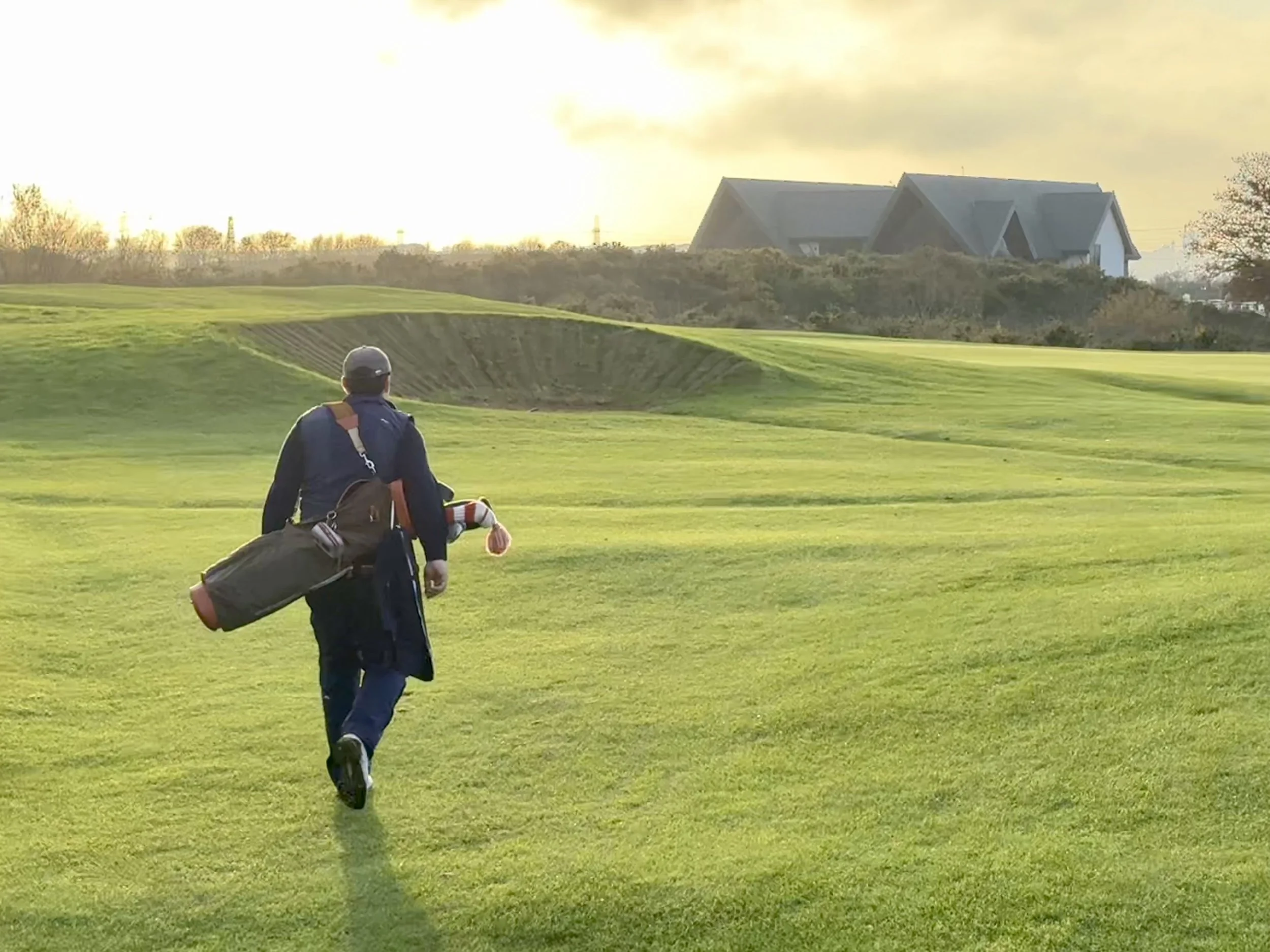 A golfer walking across a lush green golf course with a golf bag over his shoulder, heading towards the green as the sun sets, with houses in the distance.