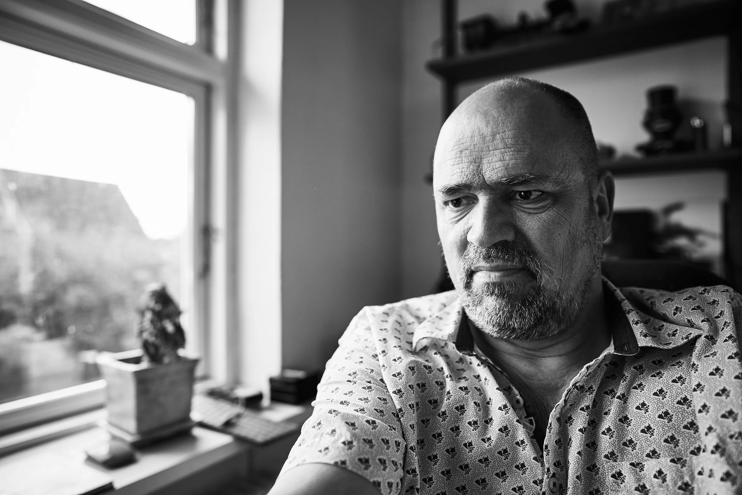 Black and white photo of a man with a beard and a shaved head sitting indoors near a window with sunlight. Shelves with objects in the background.