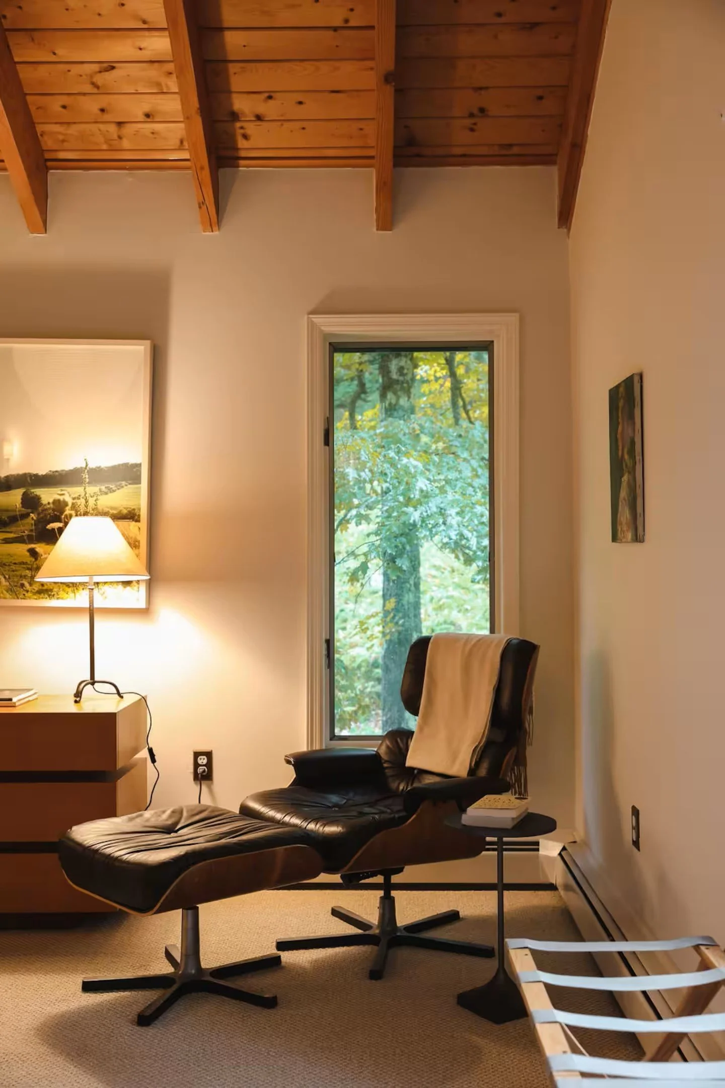 Cozy room with wooden ceiling, leather lounge chair, small side table, floor lamp on a cabinet, and a large window with forest view.