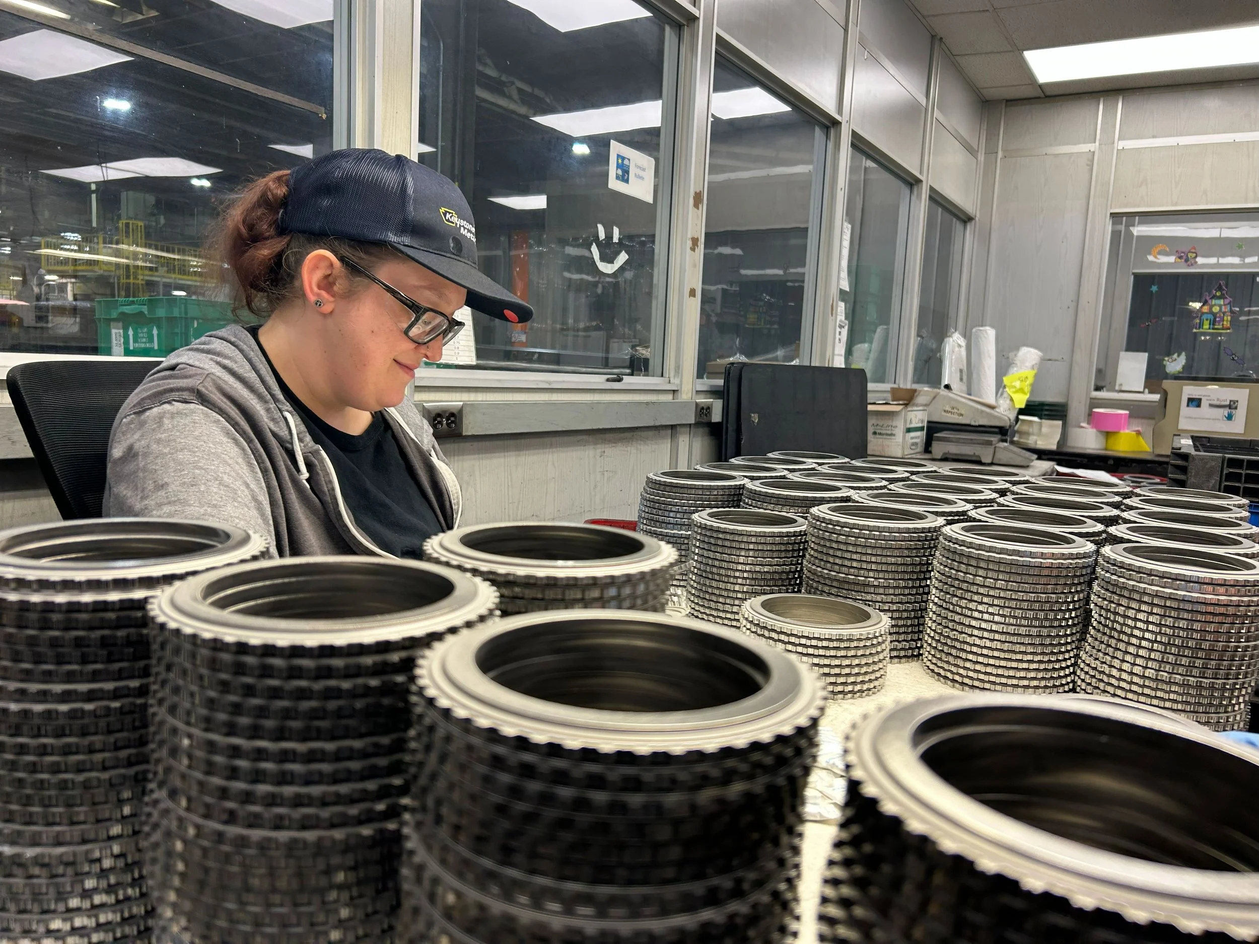 Person wearing glasses, a black cap, and gray hoodie working with stacked metal circular components on a table in an industrial or warehouse environment.