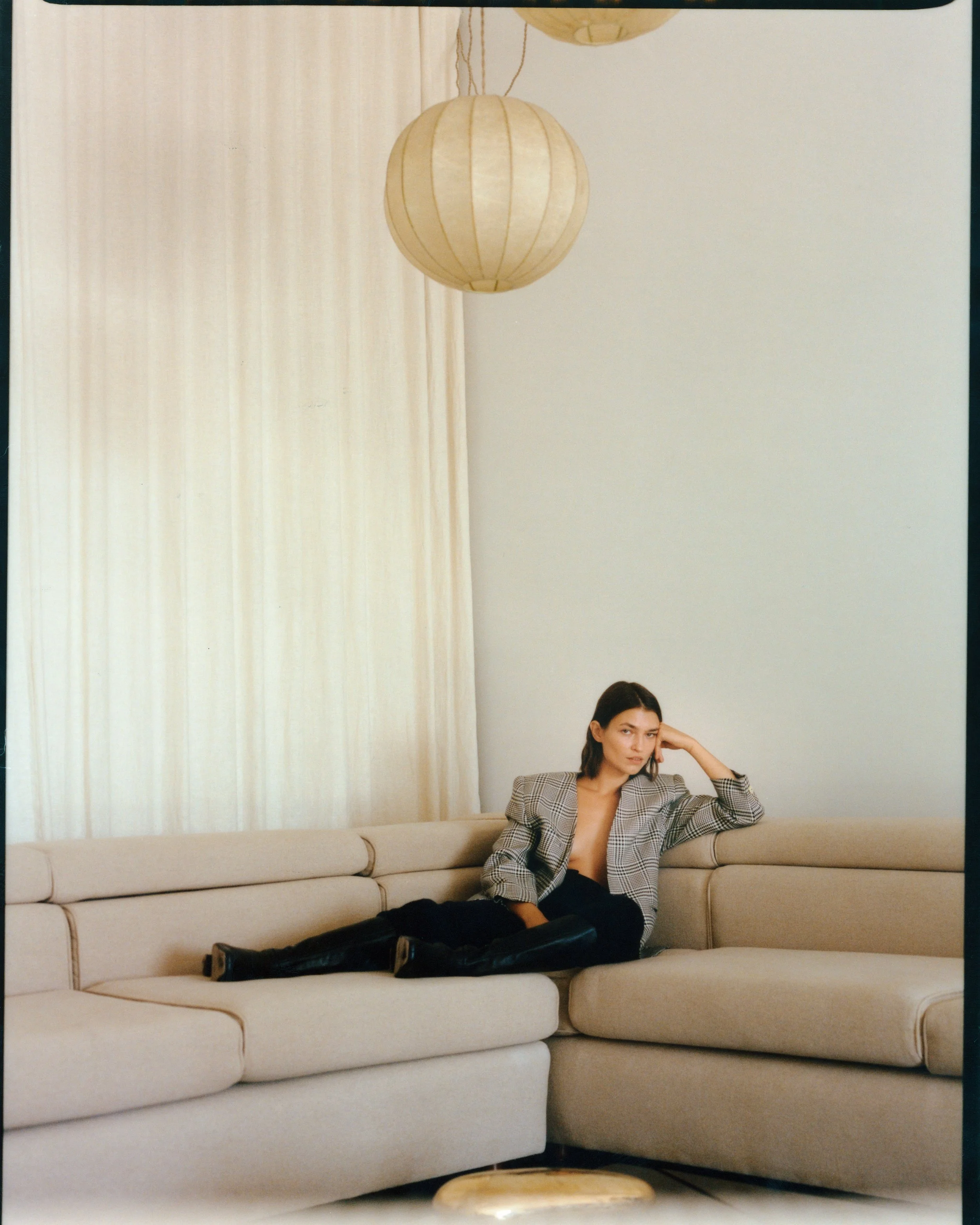 A woman reclining on a beige sectional sofa in a minimalist room with cream-colored curtains and a round paper lantern hanging from the ceiling.