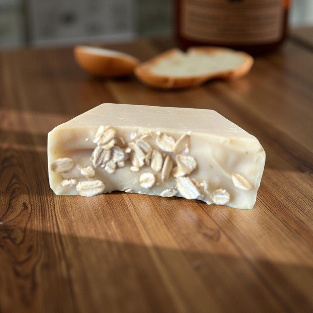 A bar of white soap with oats embedded in it, placed on a wooden surface with a blurred background that includes a dish and a container.