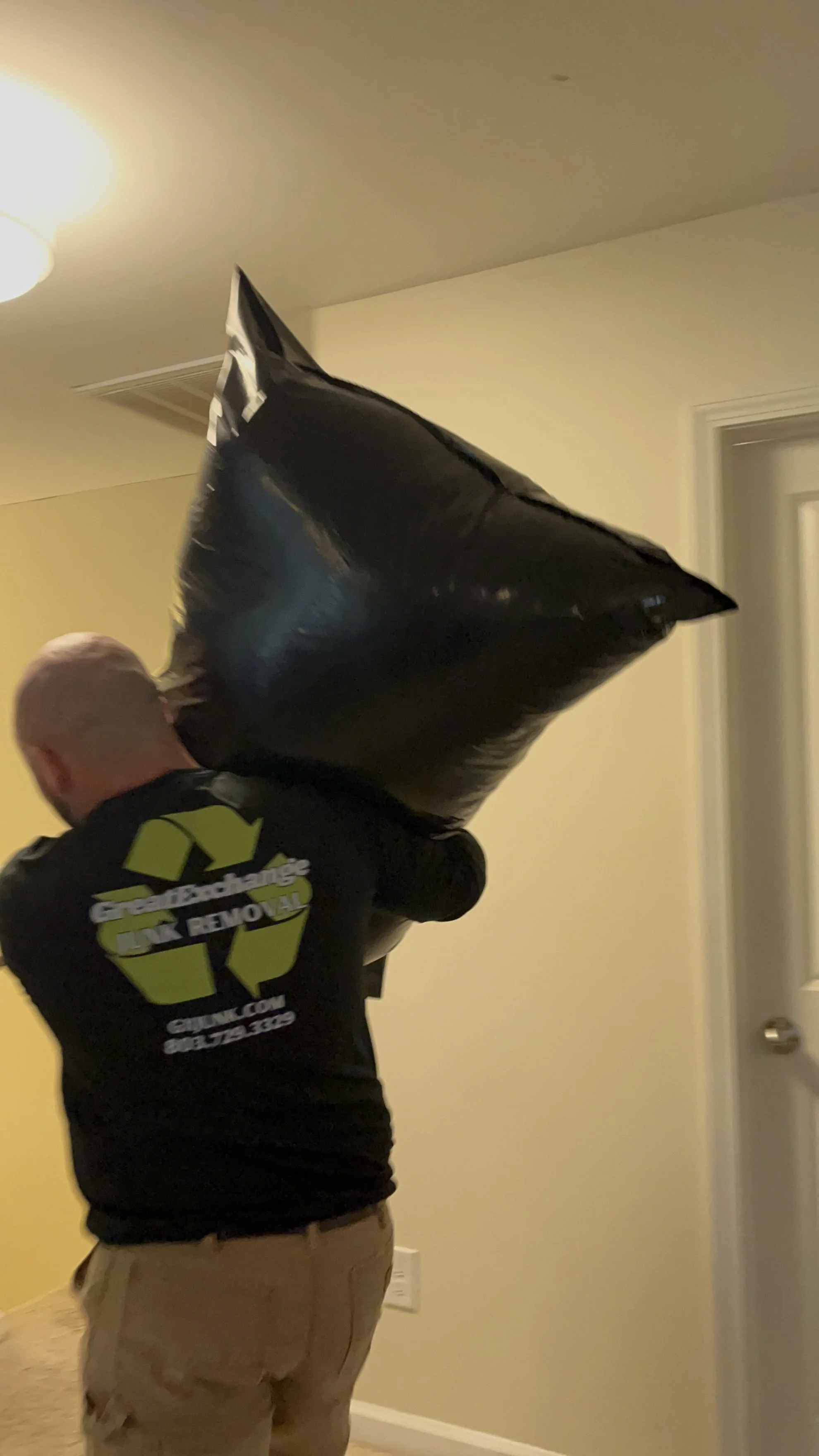 Man holding a black trash bag that has a Great Exchange Junk Removal logo on the back of his shirt, in a home.