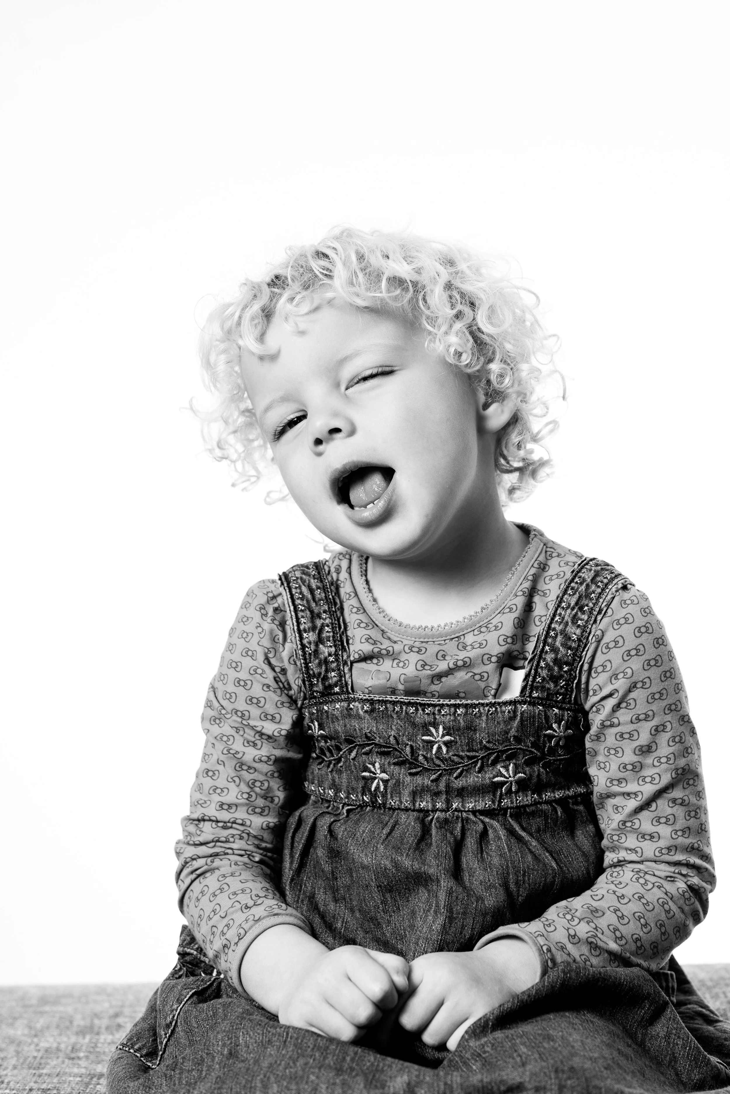 Sort-hvid foto af en ung dreng med krøllet hår, der sidder ned og ser fjollet ud med munden åben og øjne halvt lukkede, iført en langærmet skjorte med mønster og en denimkjole.