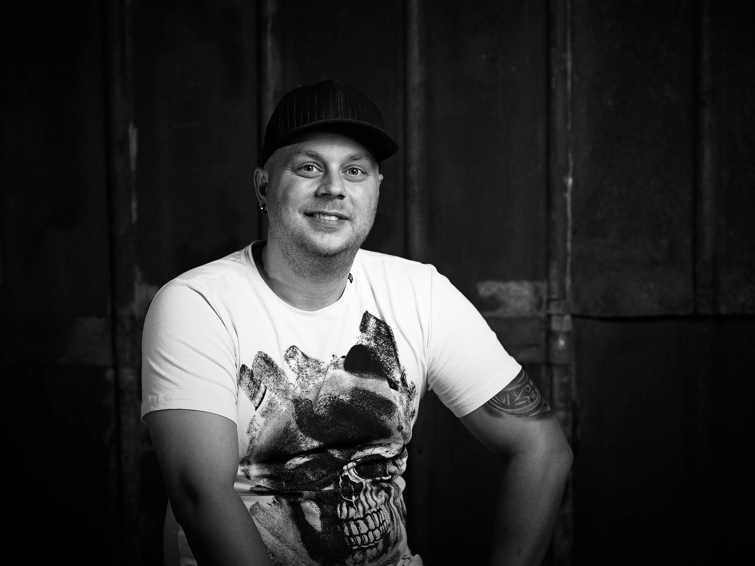 Sort-hvid portrætfoto af en mand med kort hår, ørestikker, tatovering på armen, iført t-shirt med dødningehovedmotiv, siddende foran en mørk baggrund.