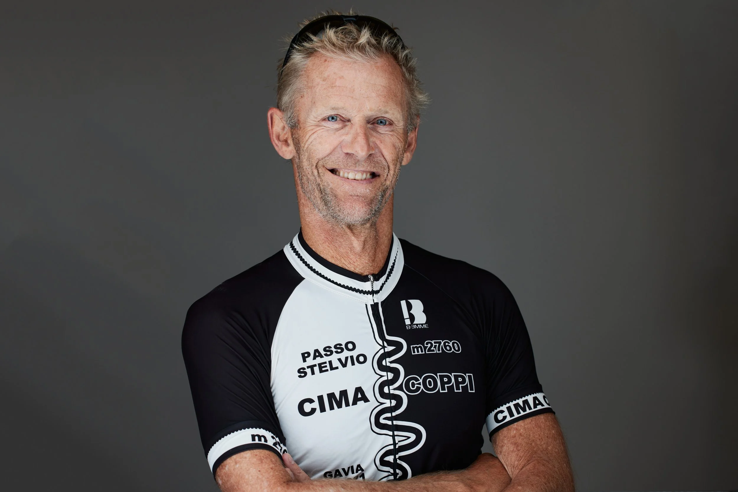 En ældre mand med blondt hår og blå øjne, iført en sort og hvid cykeltrøje med flere sponsorlogoer, smiler til kameraet mod en grå baggrund.