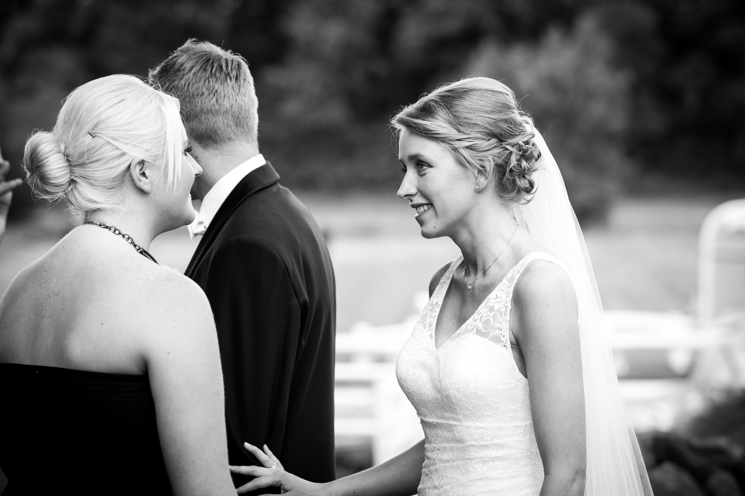 To kvinder ser på hinanden og smiler under et bryllup, en udendørs ceremoni, i sort-hvid fotografi.