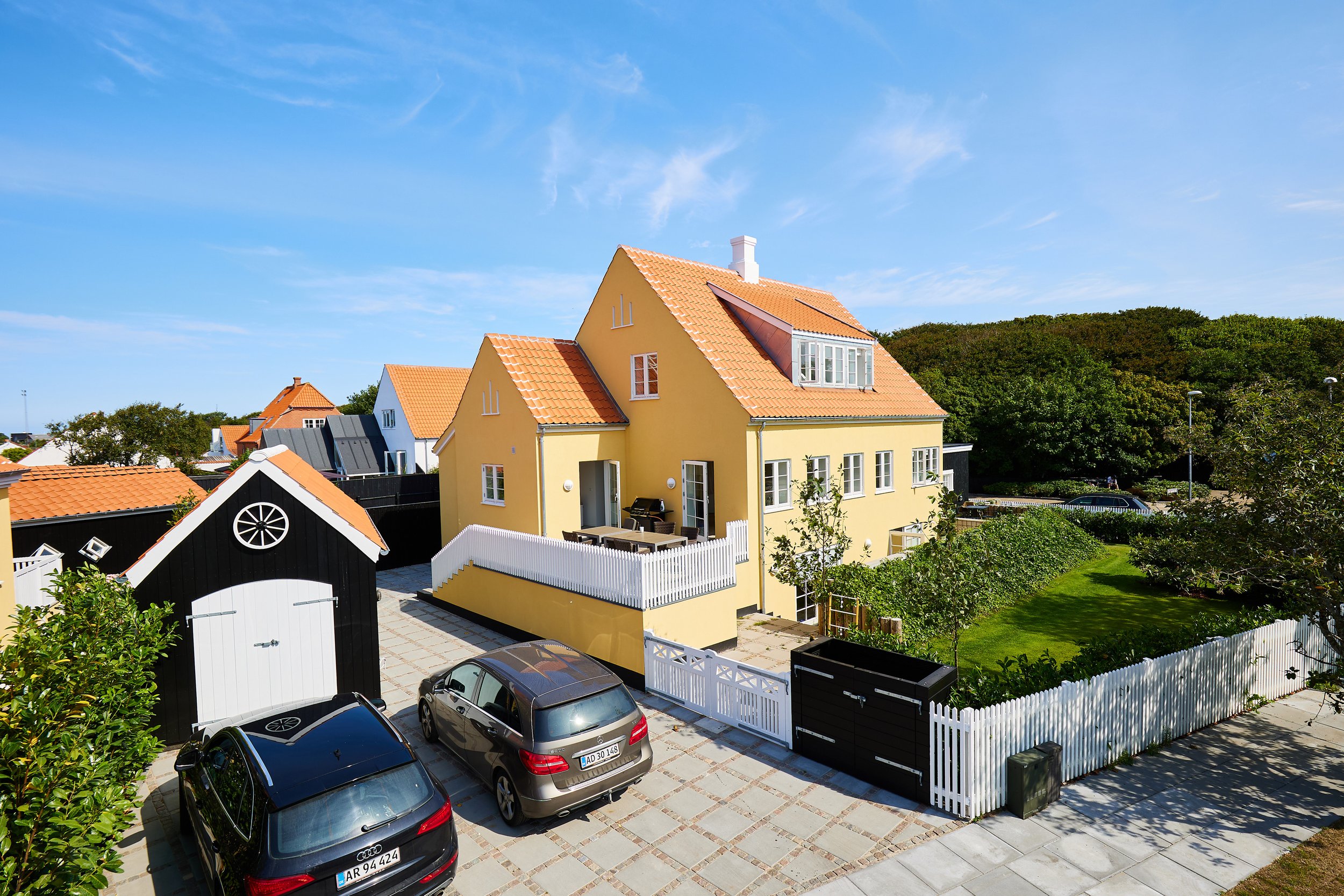 Billedet viser en gårdhave med flere farvede huse, herunder en gul med skrå tag og udskud, en sort og hvid redskabsbygning, parkeringsplads med tre biler, en hæk, et grønt område med buske og træer, en hæk og en græsplæne, med en blå himmel med få sk