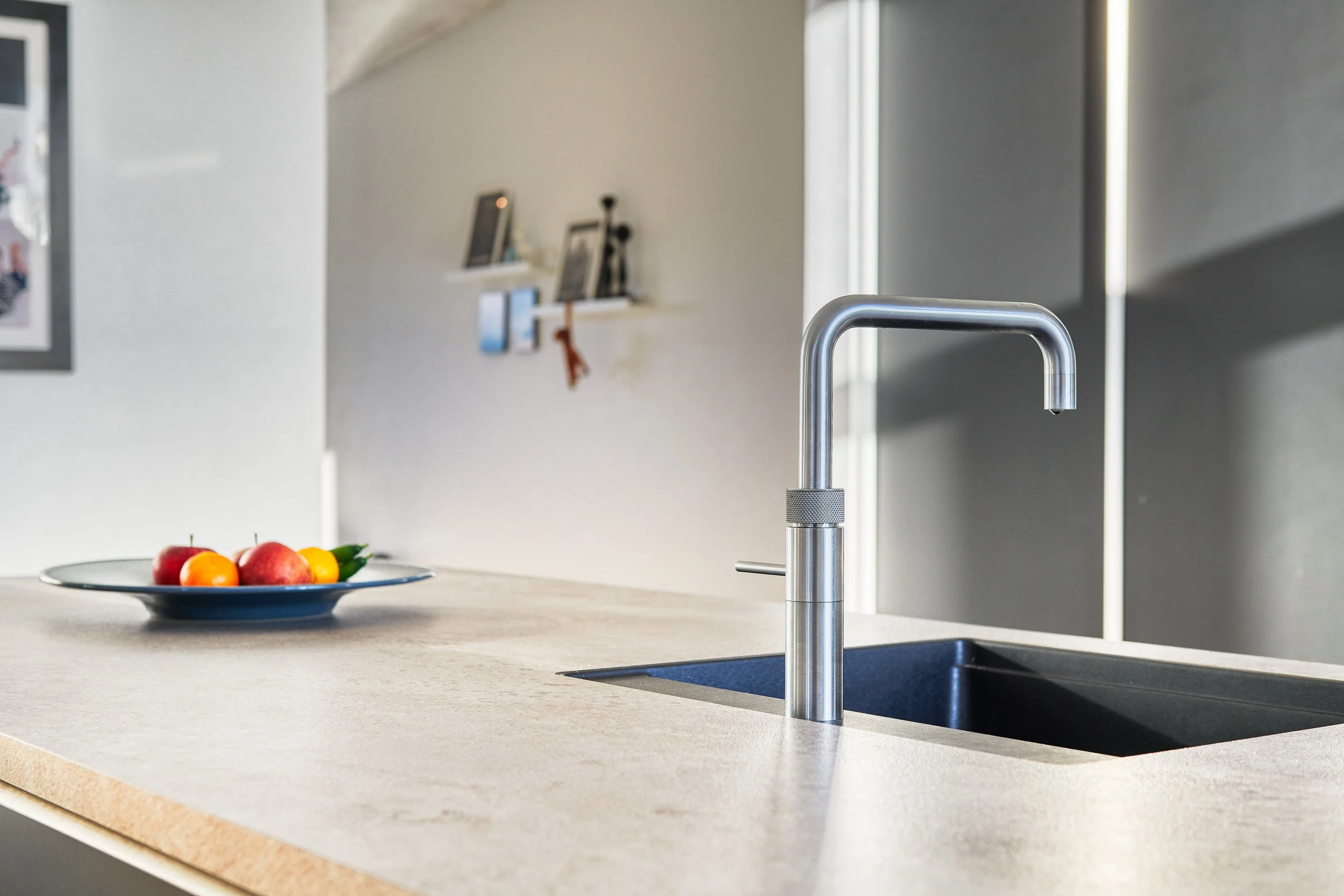 Et køkkenbord med en fyld-fad med æbler og gule stikkelsbær, en moderne vask med en krom-krystal, og en baggrund med en hvid væg med billeder og hylder.
