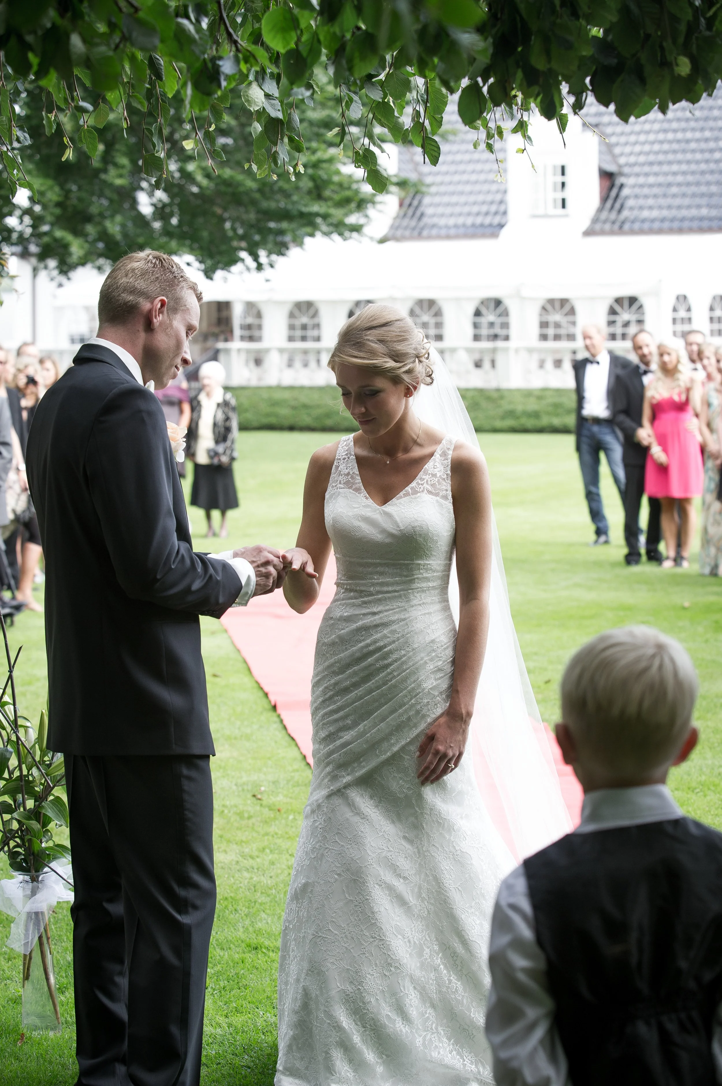 En bryllupsceremoni med en brud og en brudgom, der holder hinanden i hænderne, omgivet af venner og familie udendørs på græsset foran en hvid ferdig bygning.