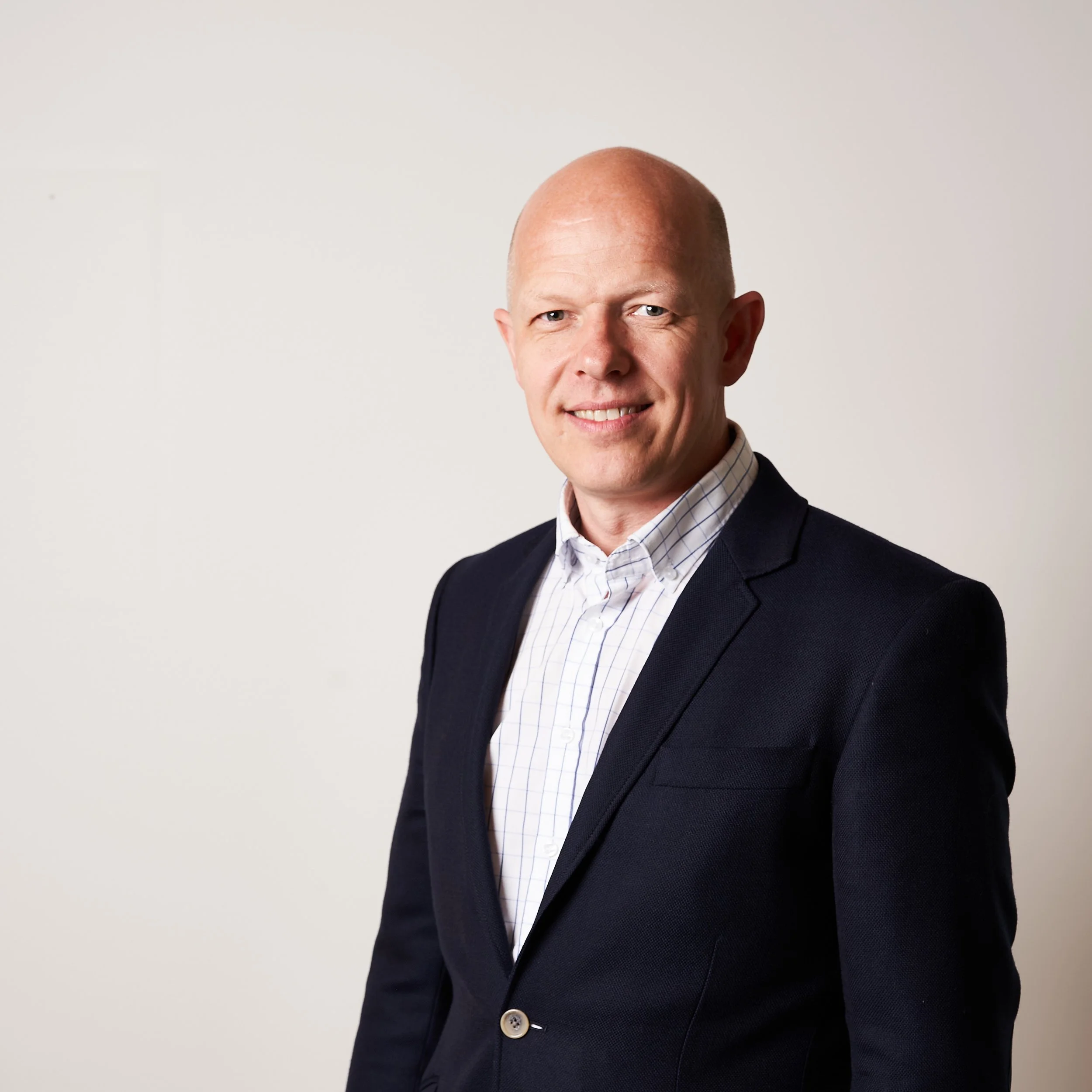 Et portræt af en mand iført en mørk blazer og en rutete skjorte, med et smil, og en helt skaldet hoved, stående mod en lys baggrund.