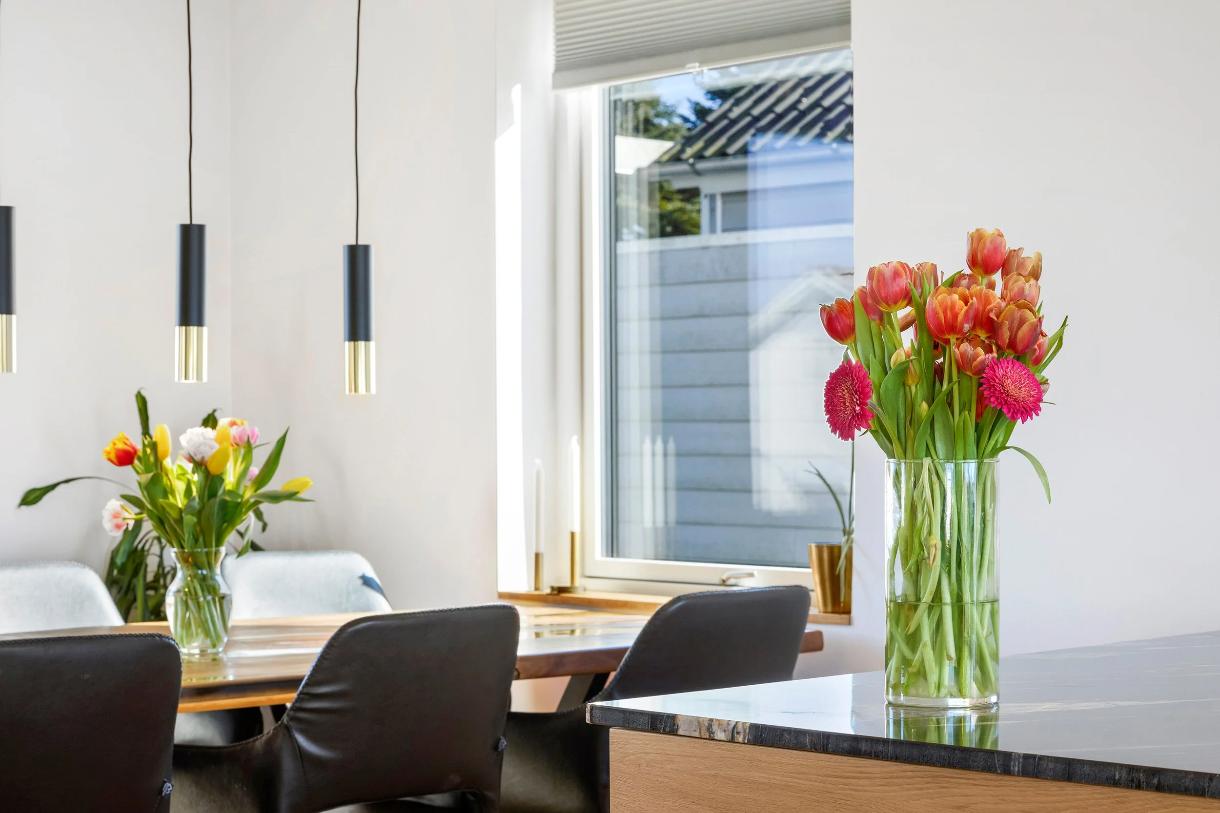 En lang glasvase med pink og orange tulipaster samt pink gerbera blomster står på et køkkenbord. I baggrunden er der et vindue, og på væggen hænger tre sorte og guldbelagte hængende lamper.