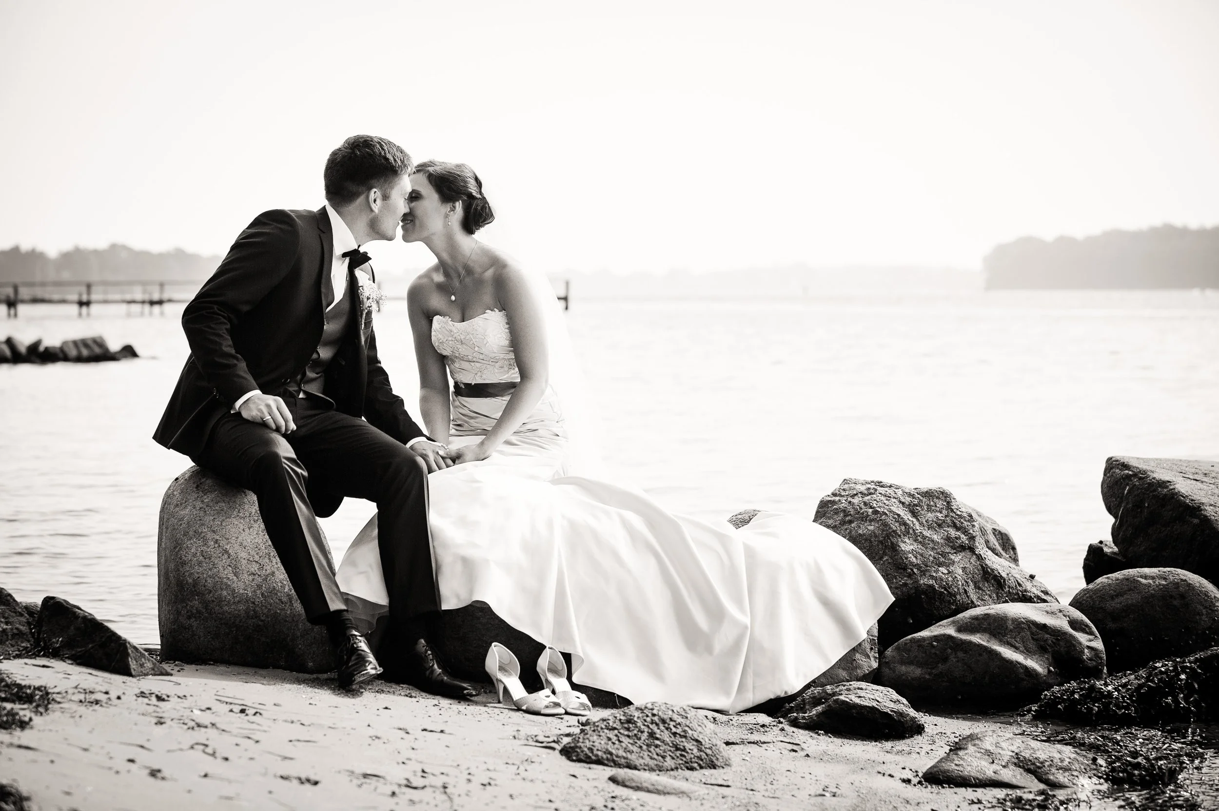 Et par i bryllupskjole og smoking sidder på store sten ved vandet og lægger ansigterne tæt sammen, ved en rå strand med sten og en pier i baggrunden, sort-hvid foto.