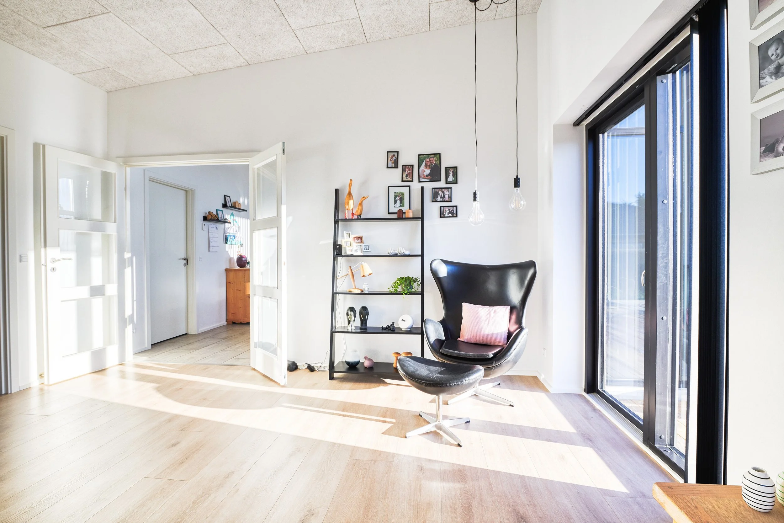 Et lyst og moderne stueområde med sort lænestol med lyserødt pudebetræk, åbne skydedøre til en stor glasdør, væg med billeder og pynt, og en hylde med dekorative genstande.