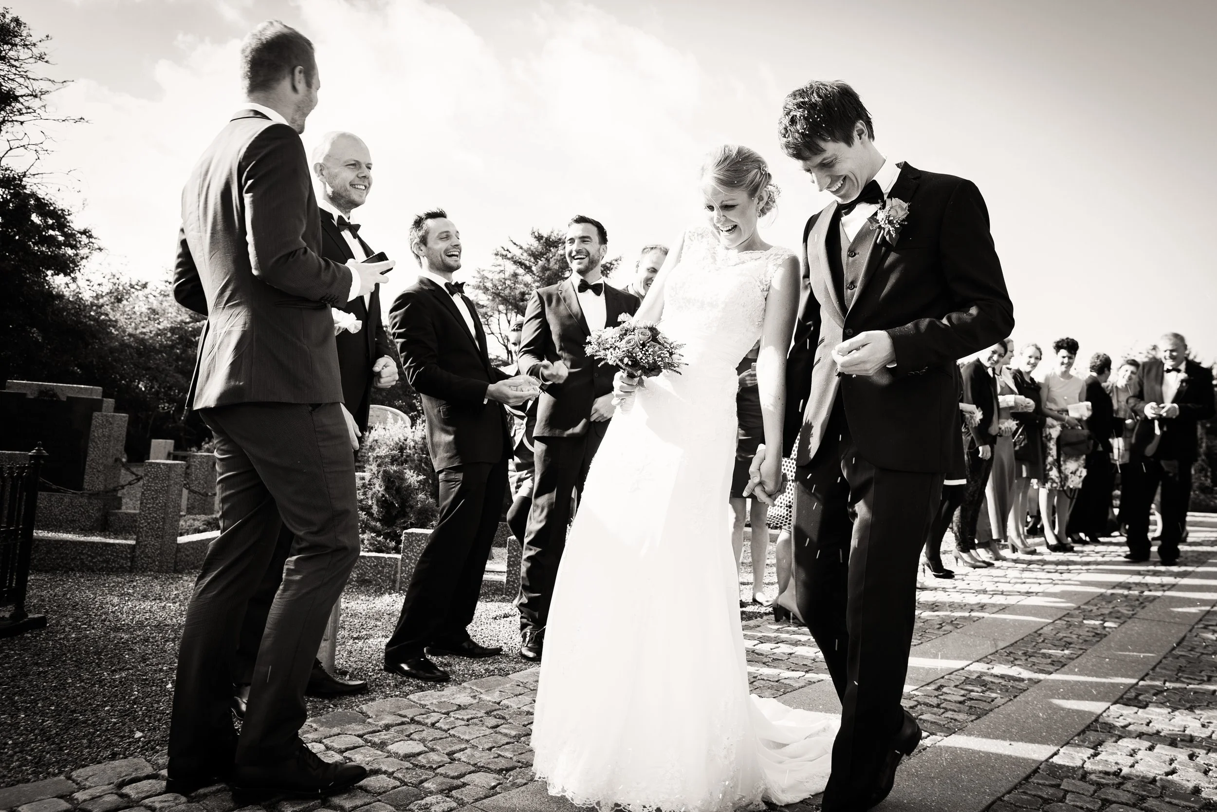 Sort-hvid foto af en bryllupsceremoni med brudeparret og venner ude i det fri. Bruden holder en blomsterbuket, og alle er klædt i formelt tøj, med mænd i smoking og kvinder i kjoler.