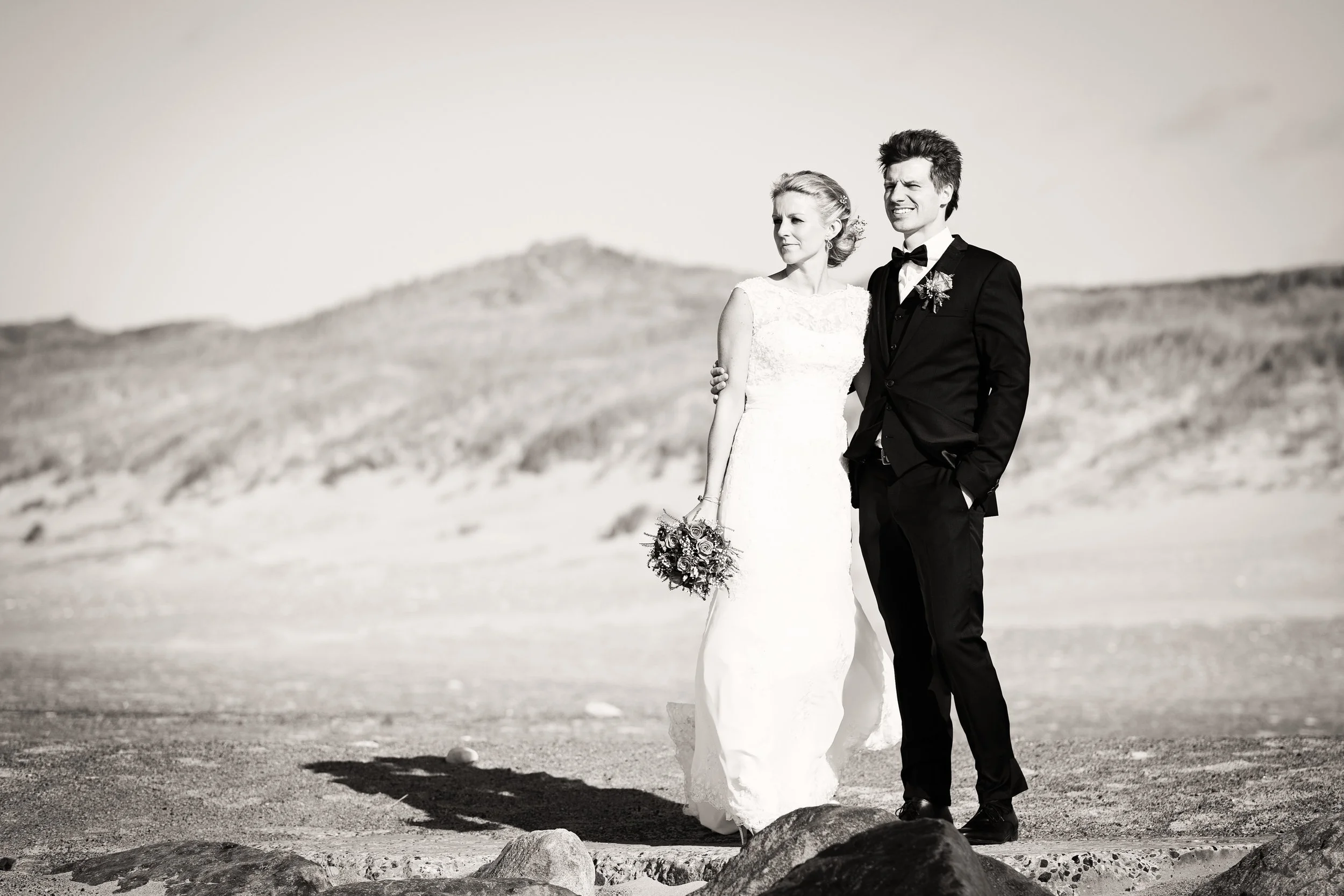 Sort-hvid foto af et bryllup med en brud i en hvid kjole og en brudgom i sort tuxedo, der står på en sten i en åben naturkreds, med bakgrund af bløde bjerge.