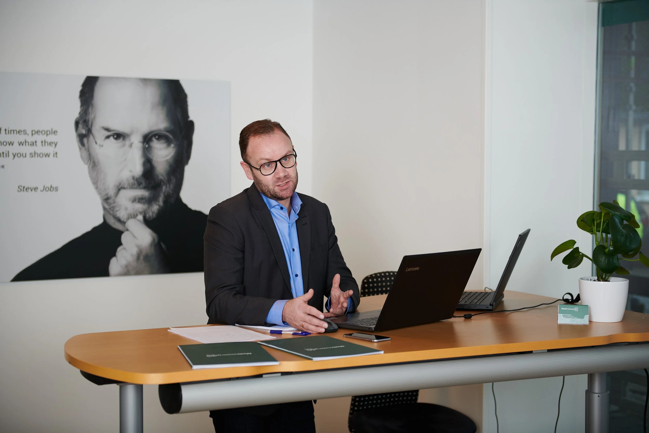 En mand med brunt hår, briller, blå skjorte og sort blazer sidder ved et træbord og taler. På bordet er en Lenovo bærbar, nogle grønne brochurer og en anden computer. Bag ham er der et stort billede af Steve Jobs og en grøn plante.