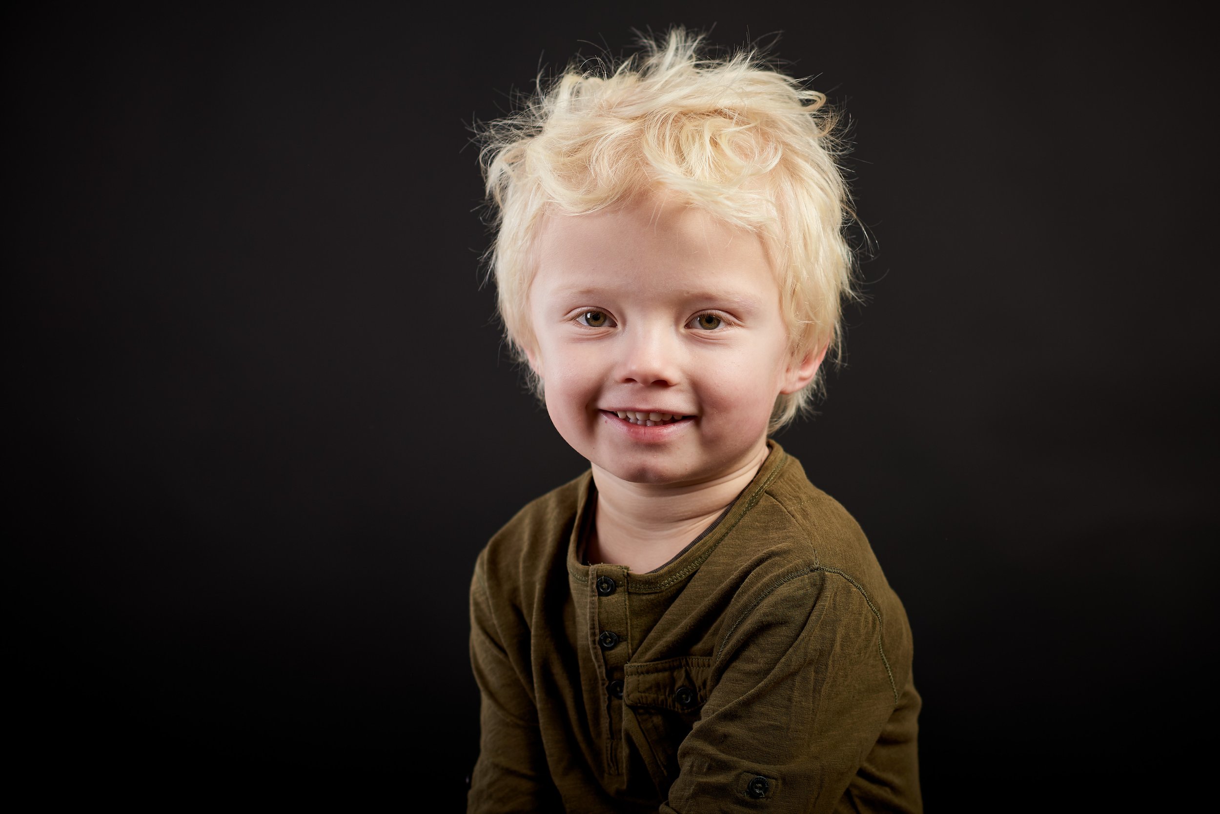 Ung dreng med lysekrøllet blondt hår, smilende og iført en grønbrun skjorte, foran sort baggrund.